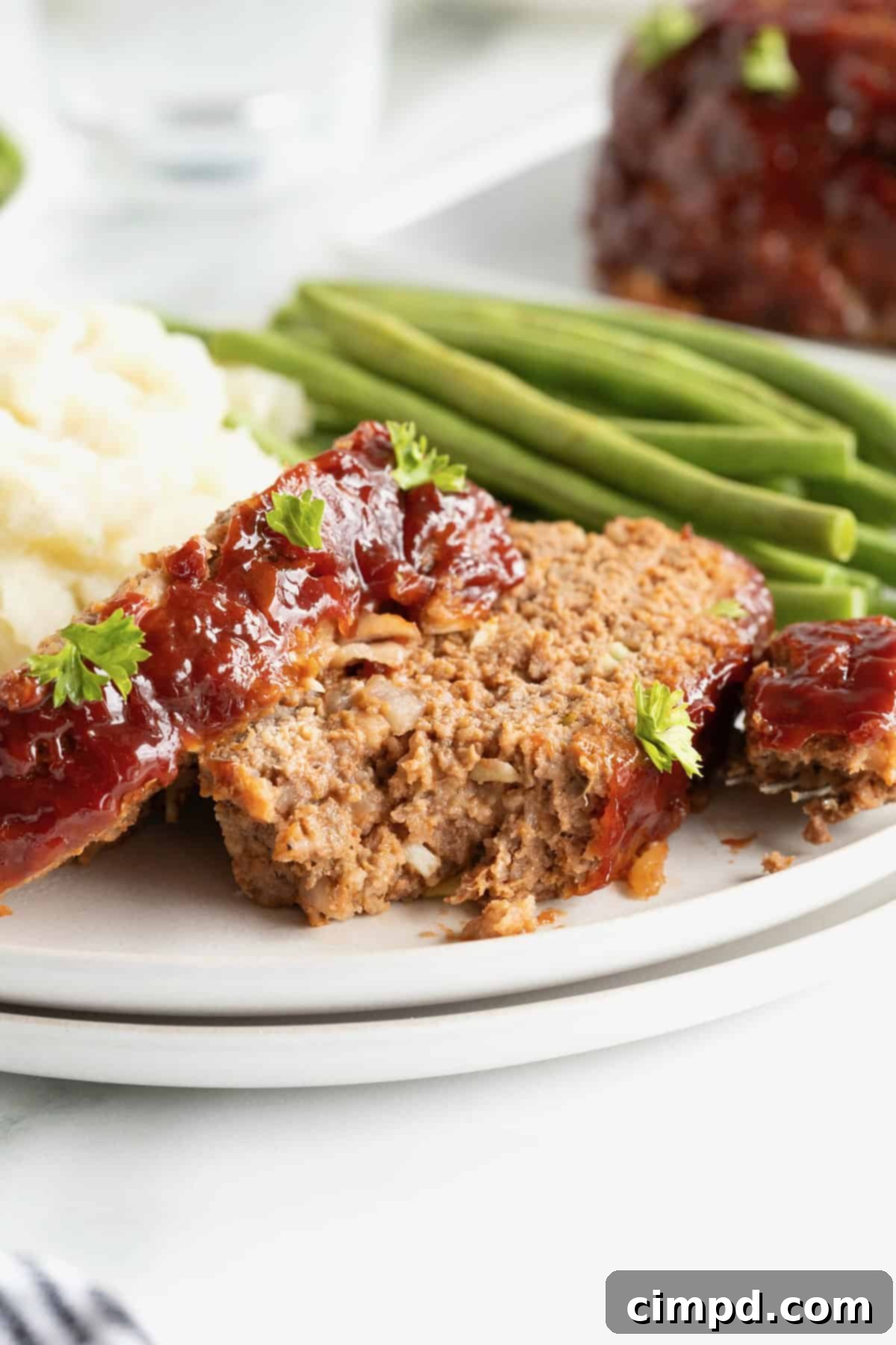 A white dinner plate showcasing two generous slices of tender meatloaf, perfectly complemented by a vibrant stack of green beans and a creamy scoop of mashed potatoes. A bite is taken from one slice, revealing the juicy interior.