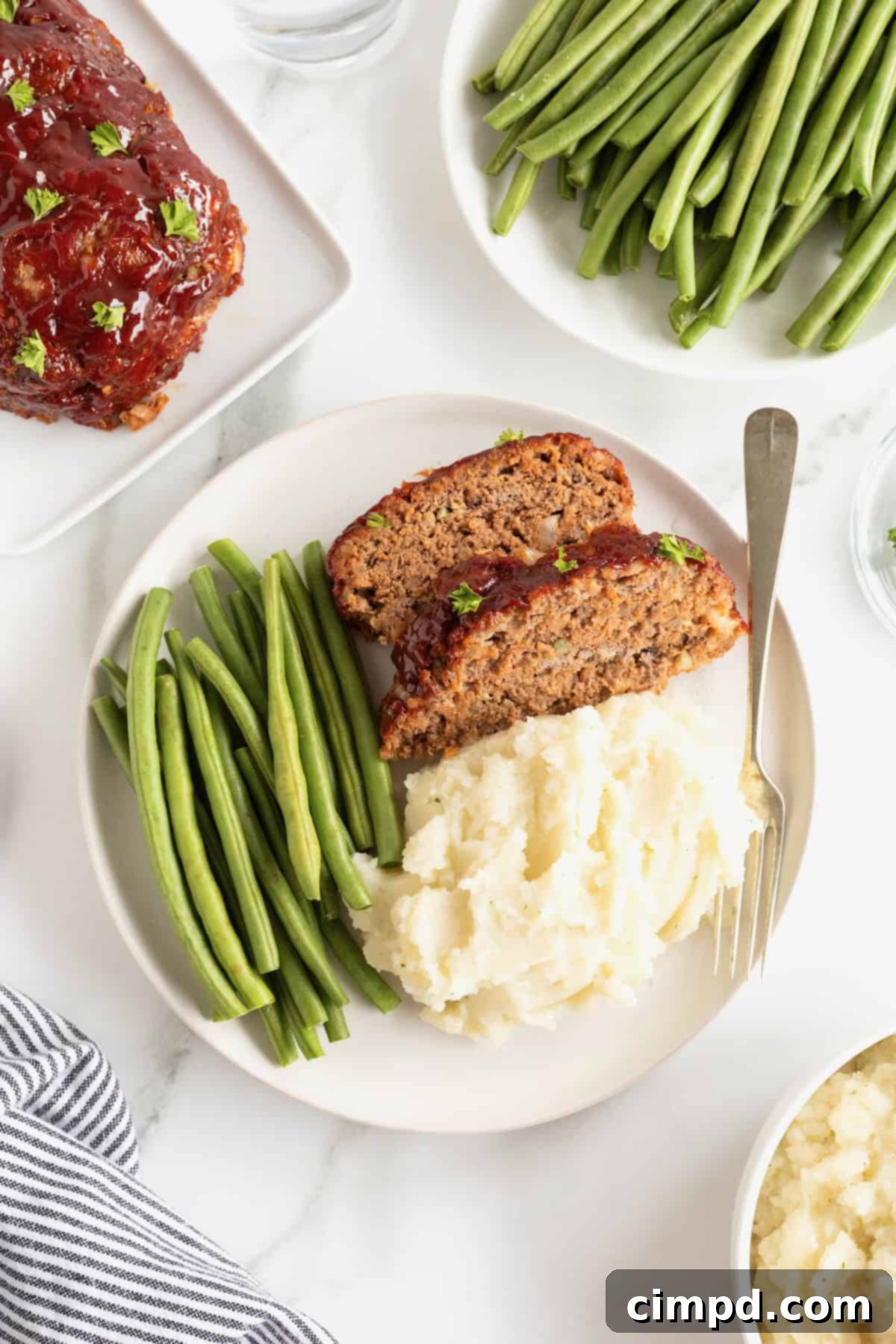 A pristine white dinner plate holding two slices of delicious meatloaf, accompanied by a neat stack of steamed green beans and a generous scoop of creamy mashed potatoes, forming a perfectly balanced meal.