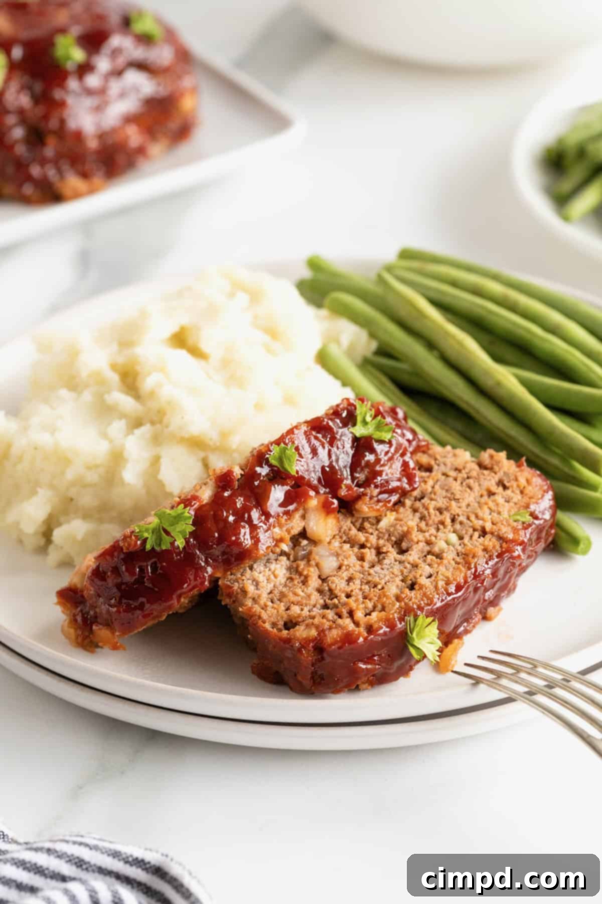 A pristine white dinner plate holding two slices of delicious meatloaf, accompanied by a neat stack of steamed green beans and a generous scoop of creamy mashed potatoes, forming a perfectly balanced meal.