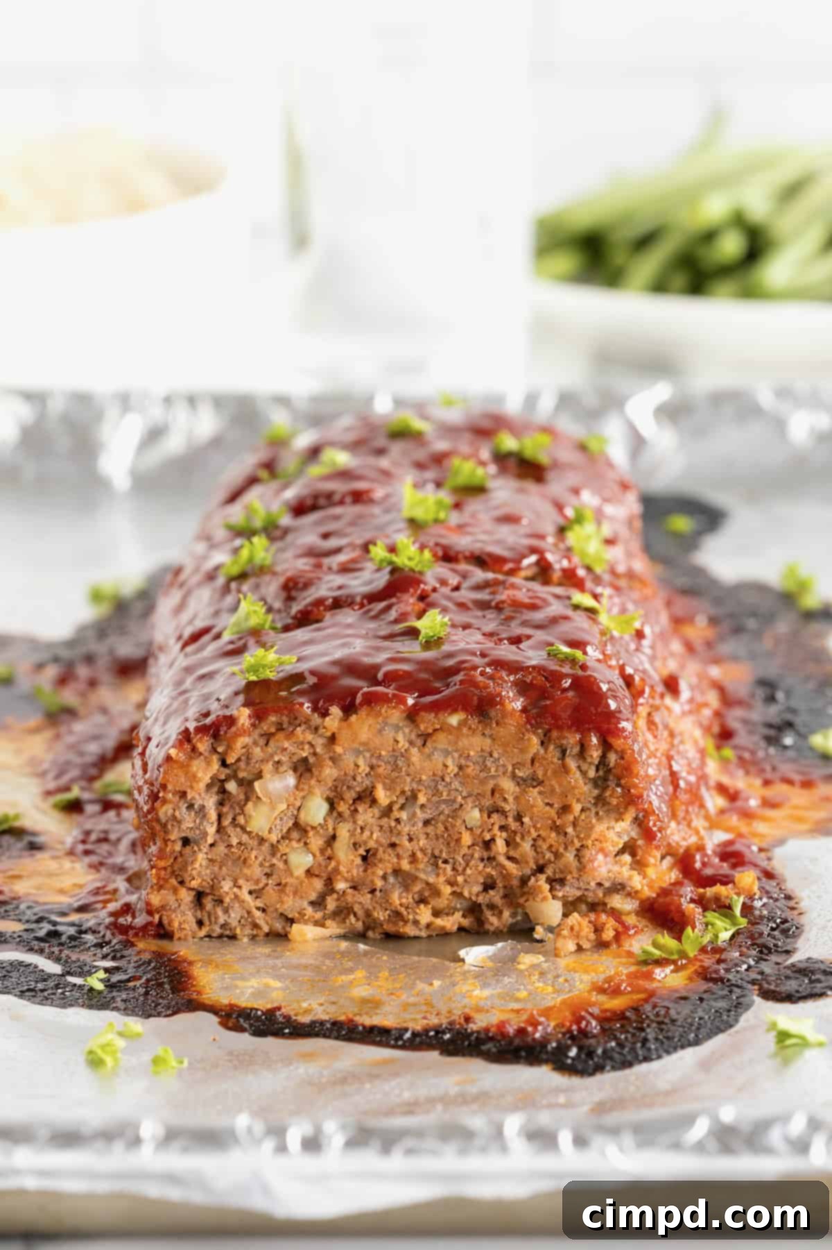 A freshly baked meatloaf, partially sliced to reveal its perfectly cooked interior, garnished with fresh parsley and resting on an aluminum foil-lined surface.