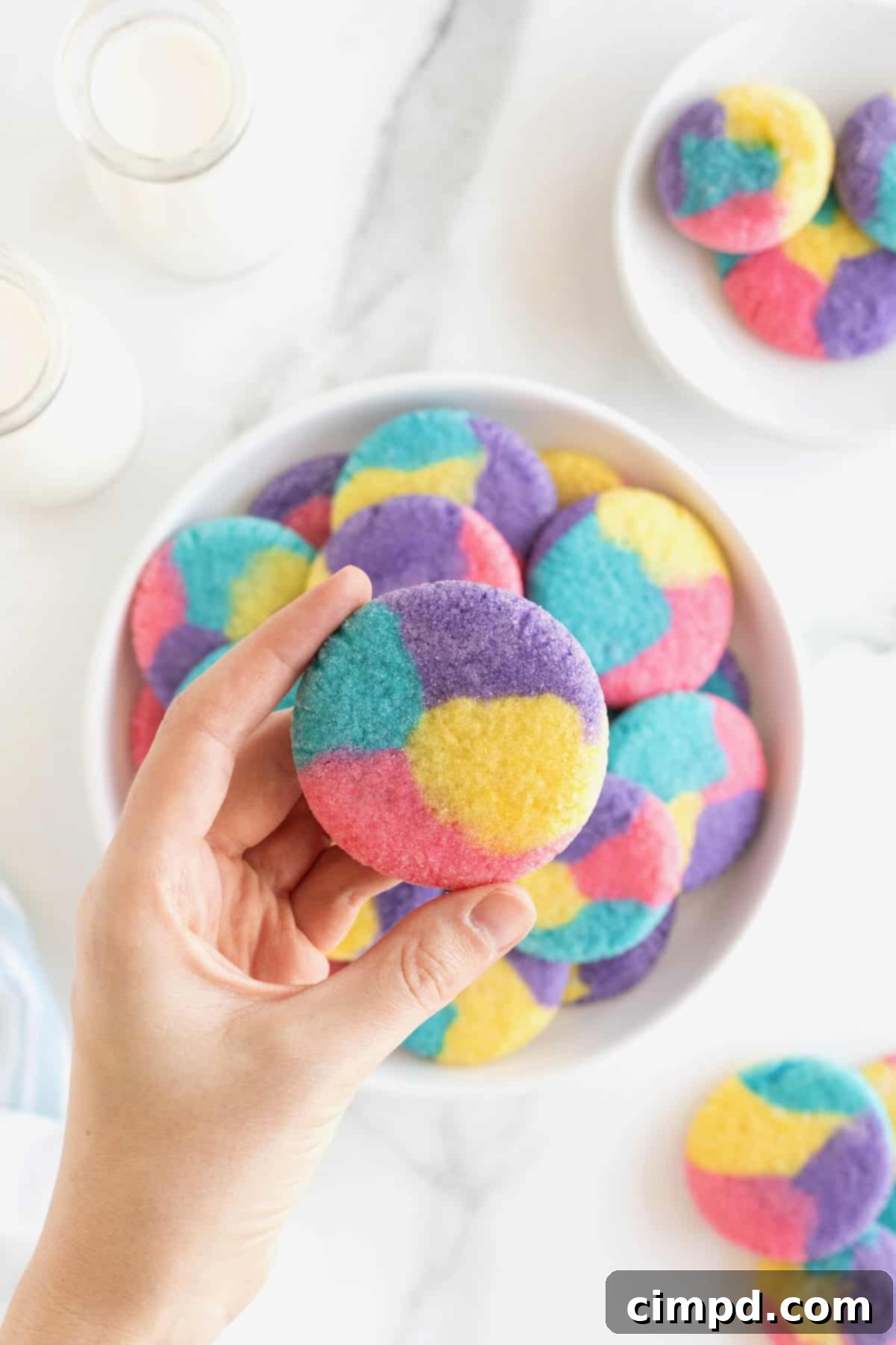 Vibrant Swirl Sugar Cookies 4 A hand holding a multi-colored sugar cookie, displaying its intricate tie-dye pattern, over a plate of freshly baked cookies.