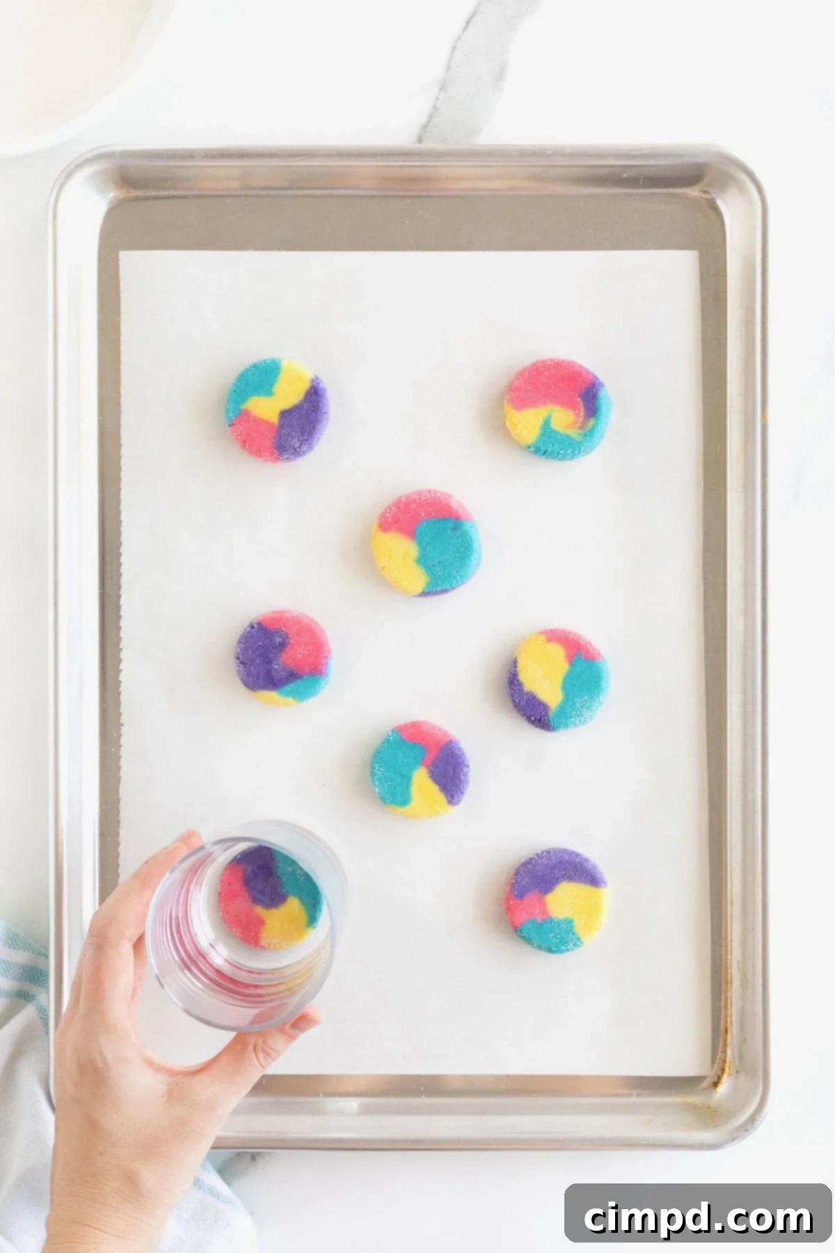 Vibrant Swirl Sugar Cookies 9 A hand gently pressing a round water glass onto a multicolored ball of cookie dough, flattening it into an even circle on the baking sheet.