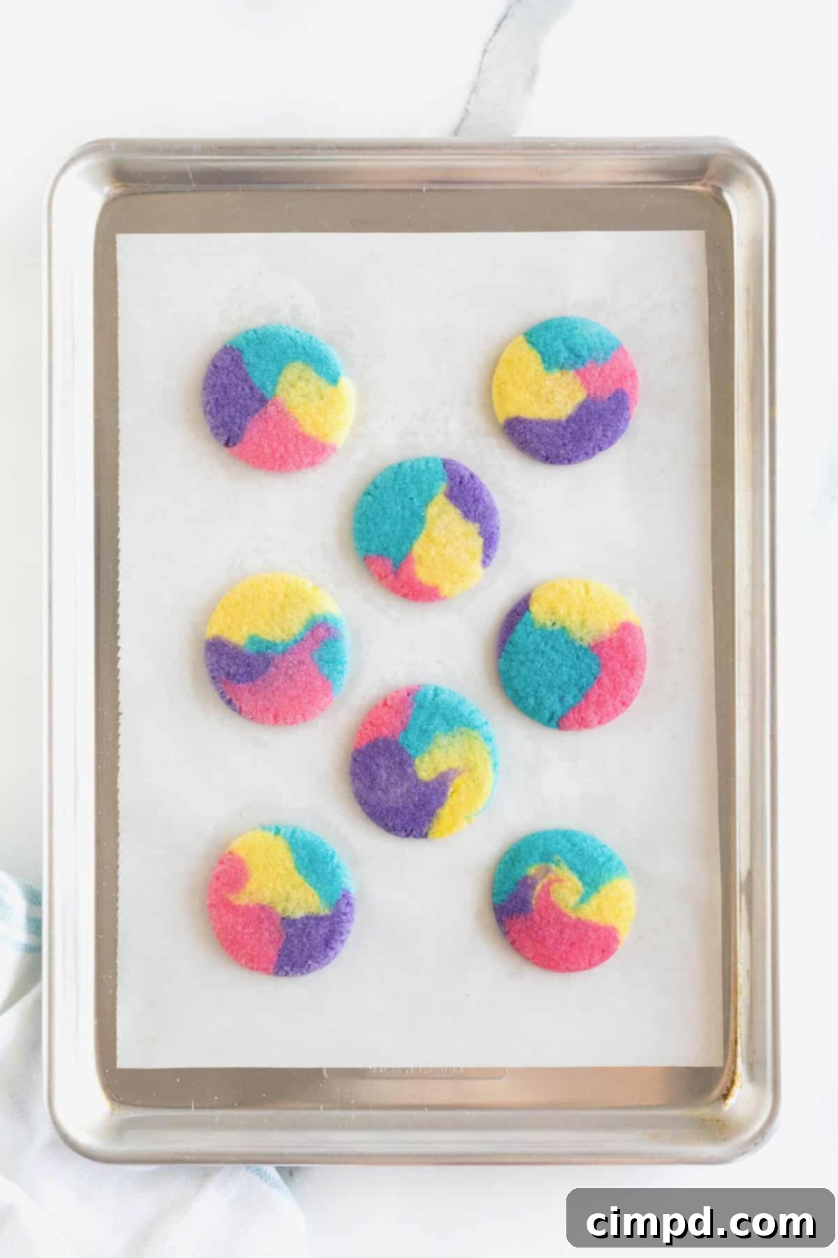 Vibrant Swirl Sugar Cookies 10 Eight fresh-baked tie-dyed sugar cookies cooling on a parchment-lined aluminum baking sheet, showing their crisp edges and vibrant colors.