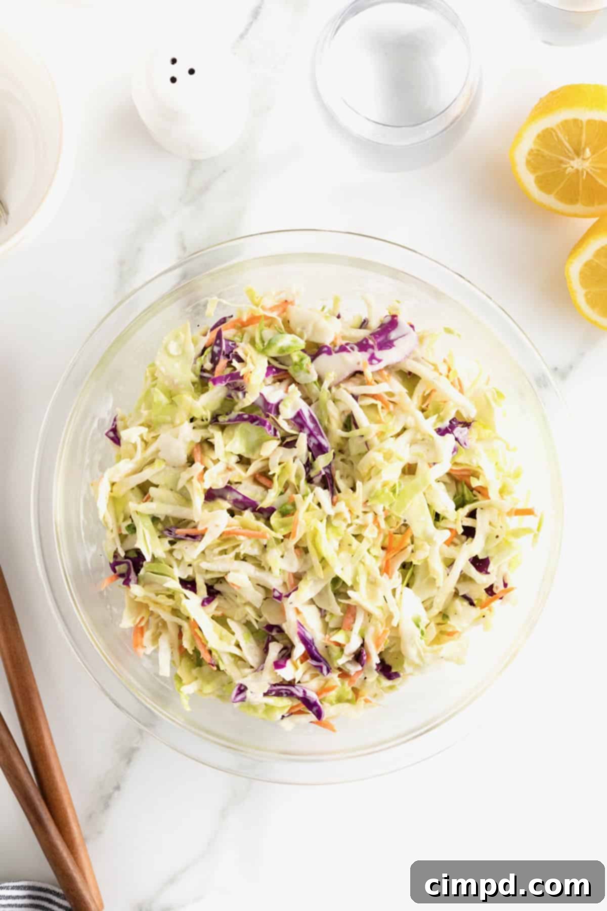 A large glass bowl of classic coleslaw on a white marble counter, with a halved lemon to its upper right.