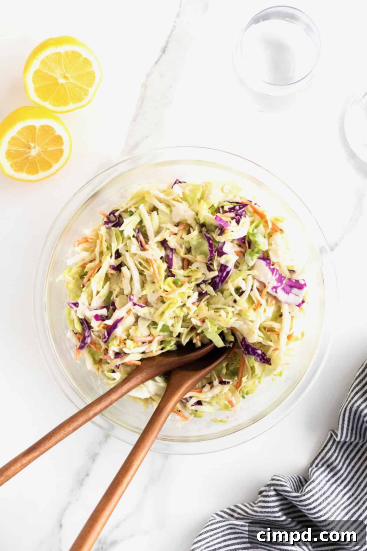 Wooden serving spoons resting in a large glass bowl of creamy coleslaw on a white marble counter, with a lemon nearby.