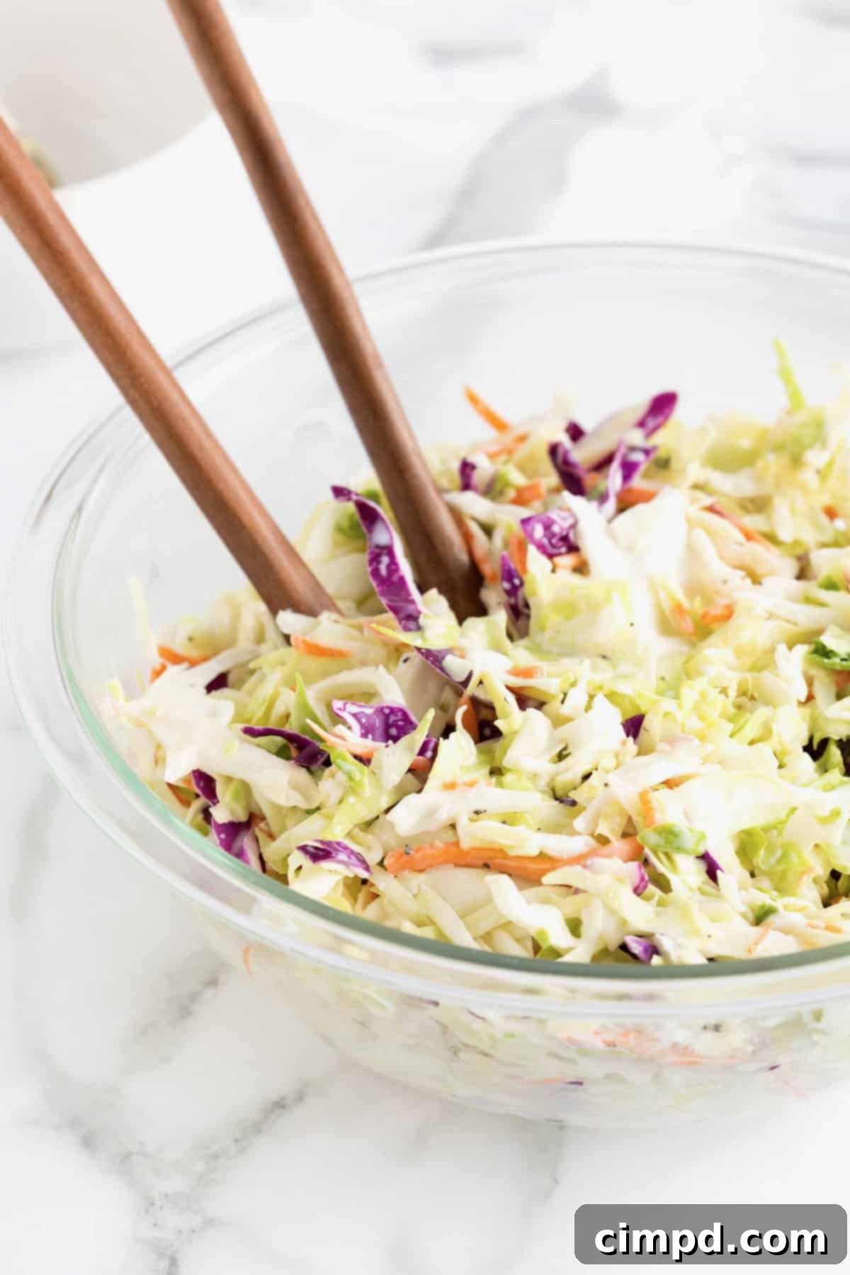 A large glass bowl of coleslaw viewed from the side, with wooden serving spoons, on a white marble counter.