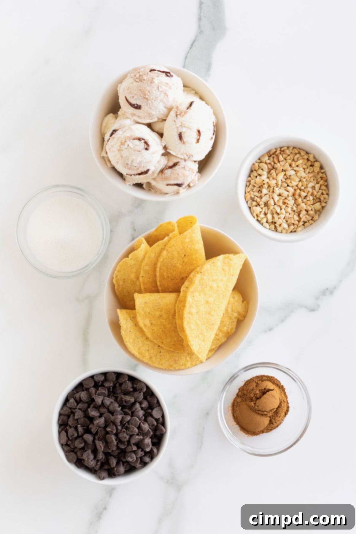 Frozen Chocolate Taco Treats 5 Various ingredients for homemade choco tacos, including mini taco shells, chocolate chips, peanuts, sugar, and cinnamon, arranged in small glass dishes on a white marble counter.
