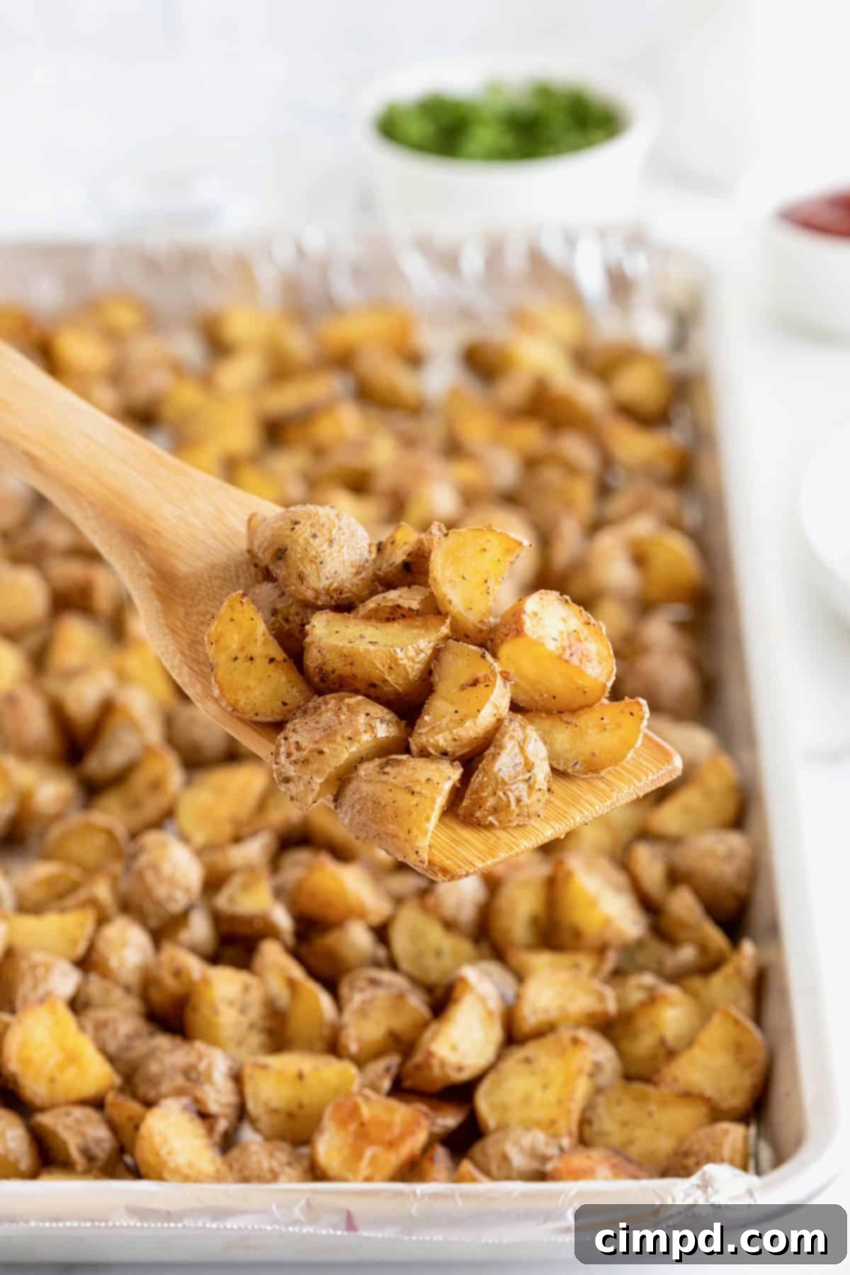 An aluminum baking sheet of oven roasted potatoes with a wooden spatula in it, illustrating a ready-to-serve or cooled batch.