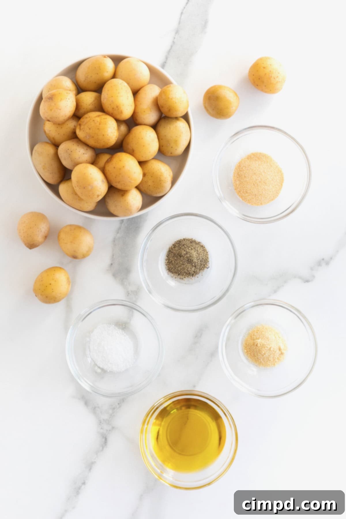 A large bowl of baby gold potatoes sits beside small glass bowls containing olive oil, salt, garlic powder, onion powder, and pepper on a white marble counter, showcasing the ingredients for Oven Roasted Crispy Potatoes.