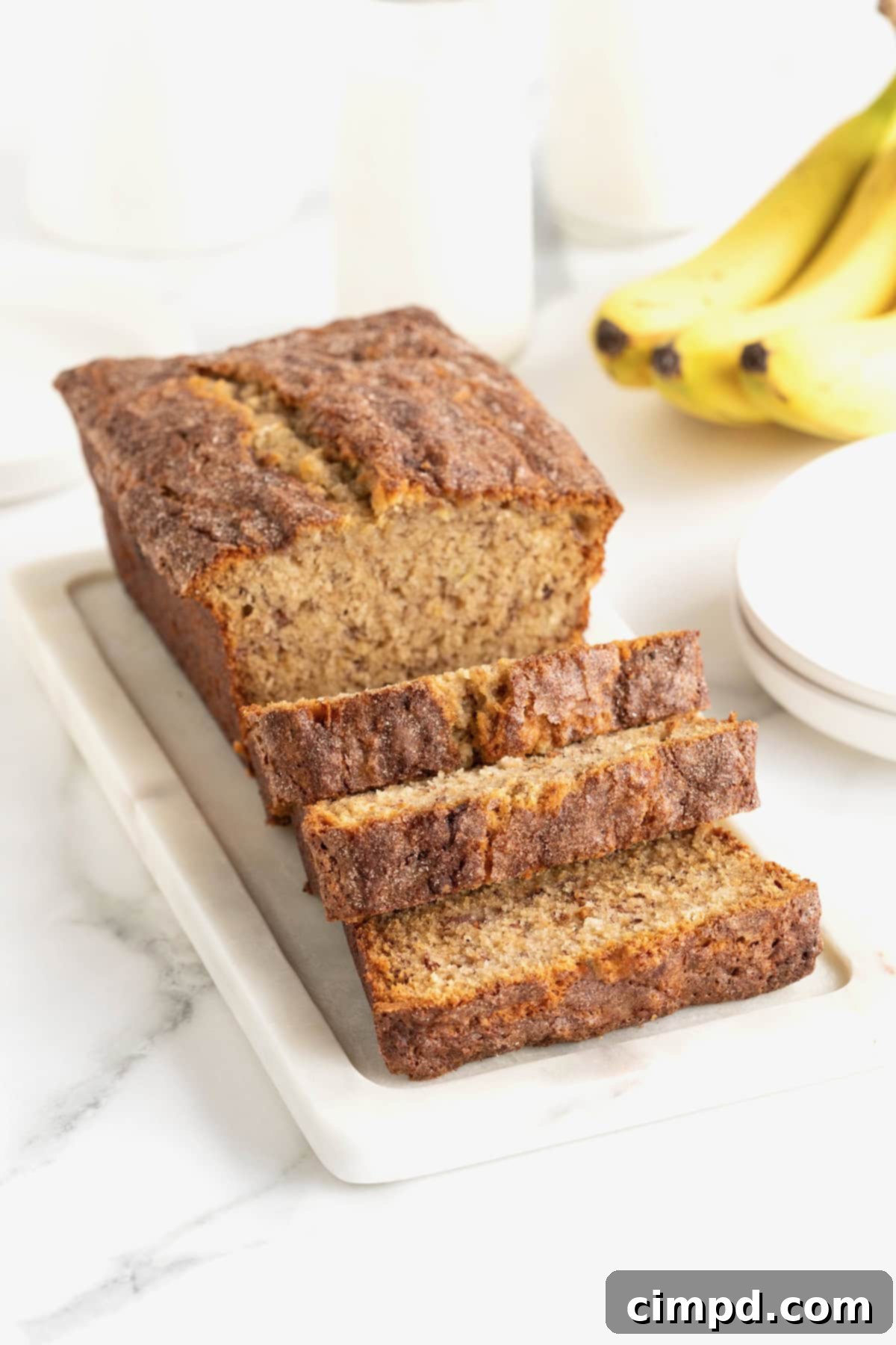 A golden-brown loaf of Cinnamon Banana Bread, generously coated in glistening cinnamon sugar, rests on a rustic white stone cutting board atop a pristine white marble counter, inviting a delicious first bite.
