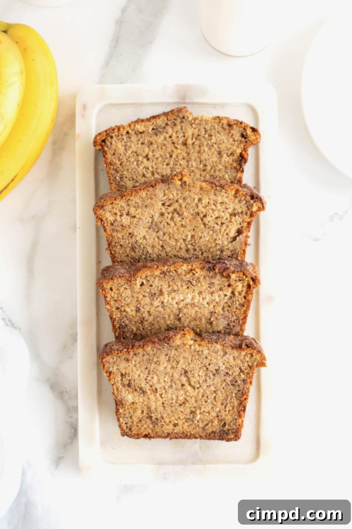 Four perfectly uniform slices of Cinnamon Banana Bread, showcasing their moist interiors and crisp cinnamon sugar crusts, are neatly arranged on a white stone cutting board, ready to be enjoyed.