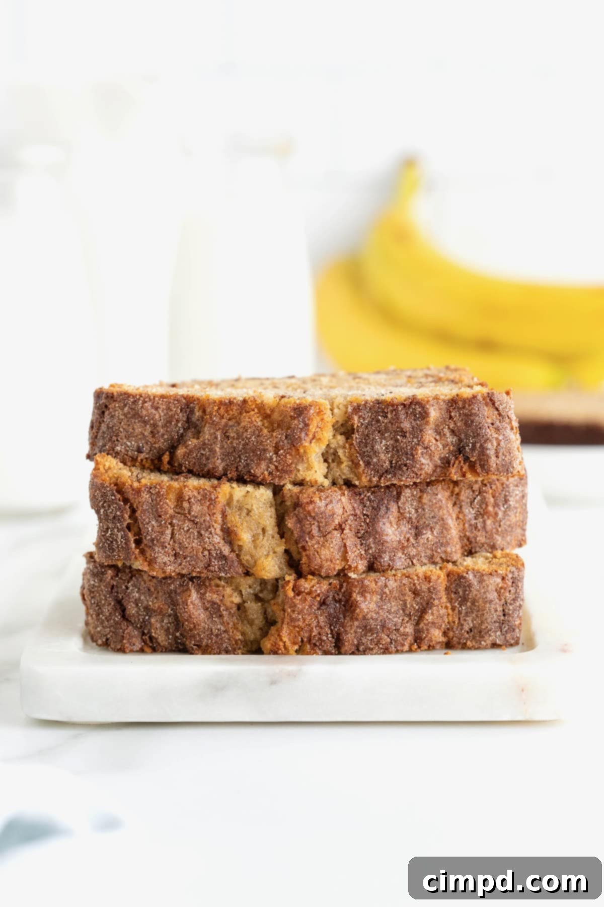 Three perfectly cut slices of Cinnamon Banana Bread, each exhibiting its inviting cinnamon sugar crust and tender, moist crumb, are carefully stacked on a rustic white stone cutting board, highlighting their appetizing appeal.