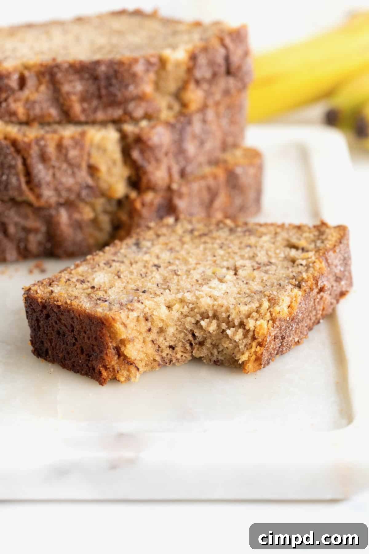 A perfectly sliced piece of Cinnamon Banana Bread, revealing its moist, tender crumb, lies on a white cutting board. The slice is beautifully dusted with cinnamon sugar, and a small bite has been taken, highlighting its inviting texture.