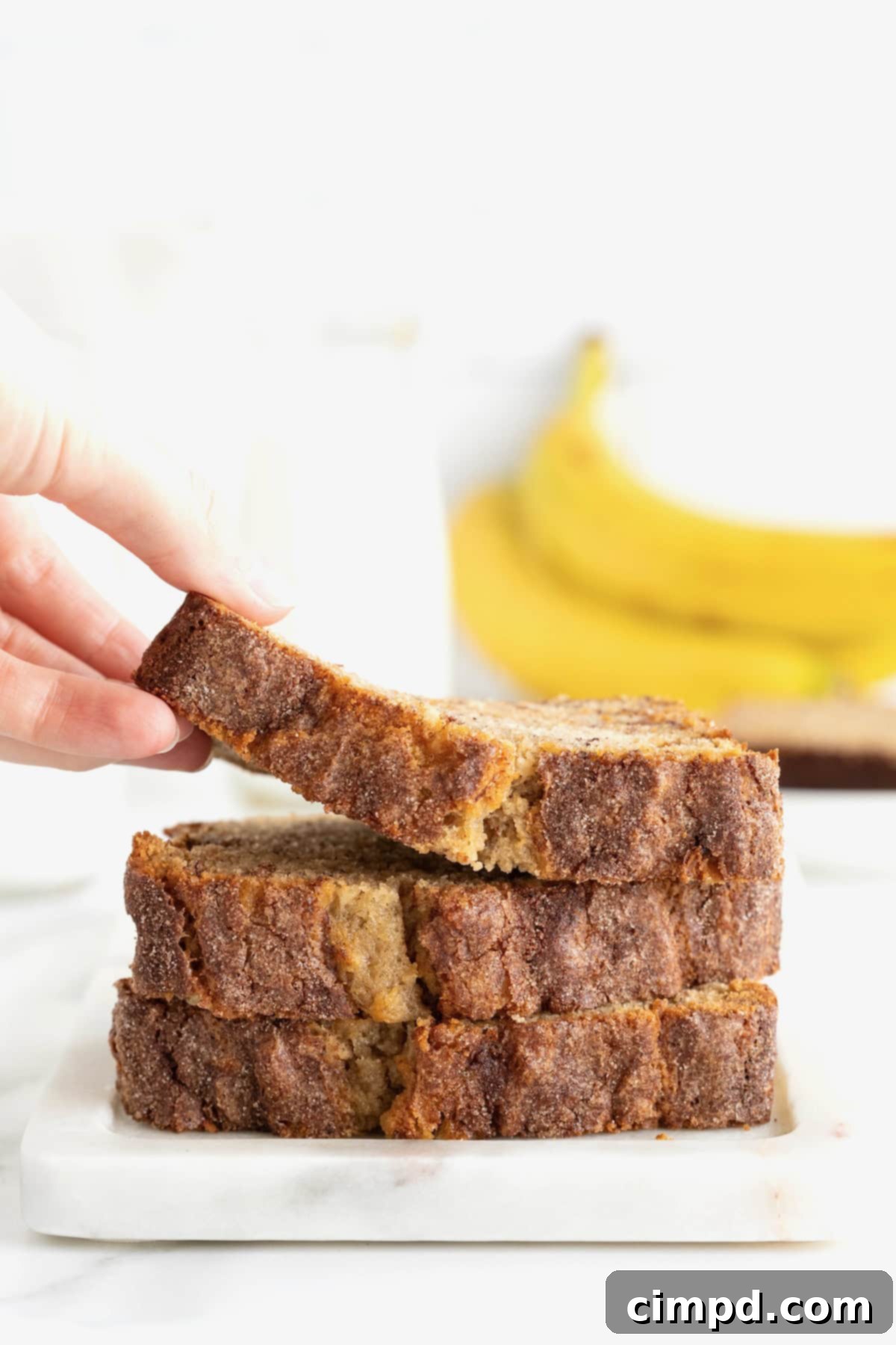 Three golden slices of Cinnamon Banana Bread, each showing its enticing cinnamon sugar coating and moist interior, are neatly stacked on a white stone cutting board. A hand gracefully lifts the top slice, hinting at its deliciousness.
