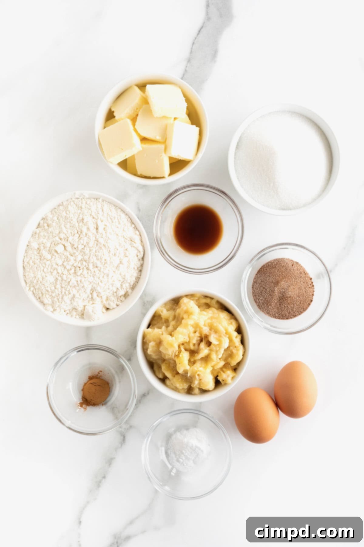 Various ingredients for making cinnamon banana bread, including butter, sugar, eggs, vanilla, mashed bananas, flour, baking soda, and cinnamon, neatly arranged in small glass dishes on a pristine white marble counter, ready for baking.
