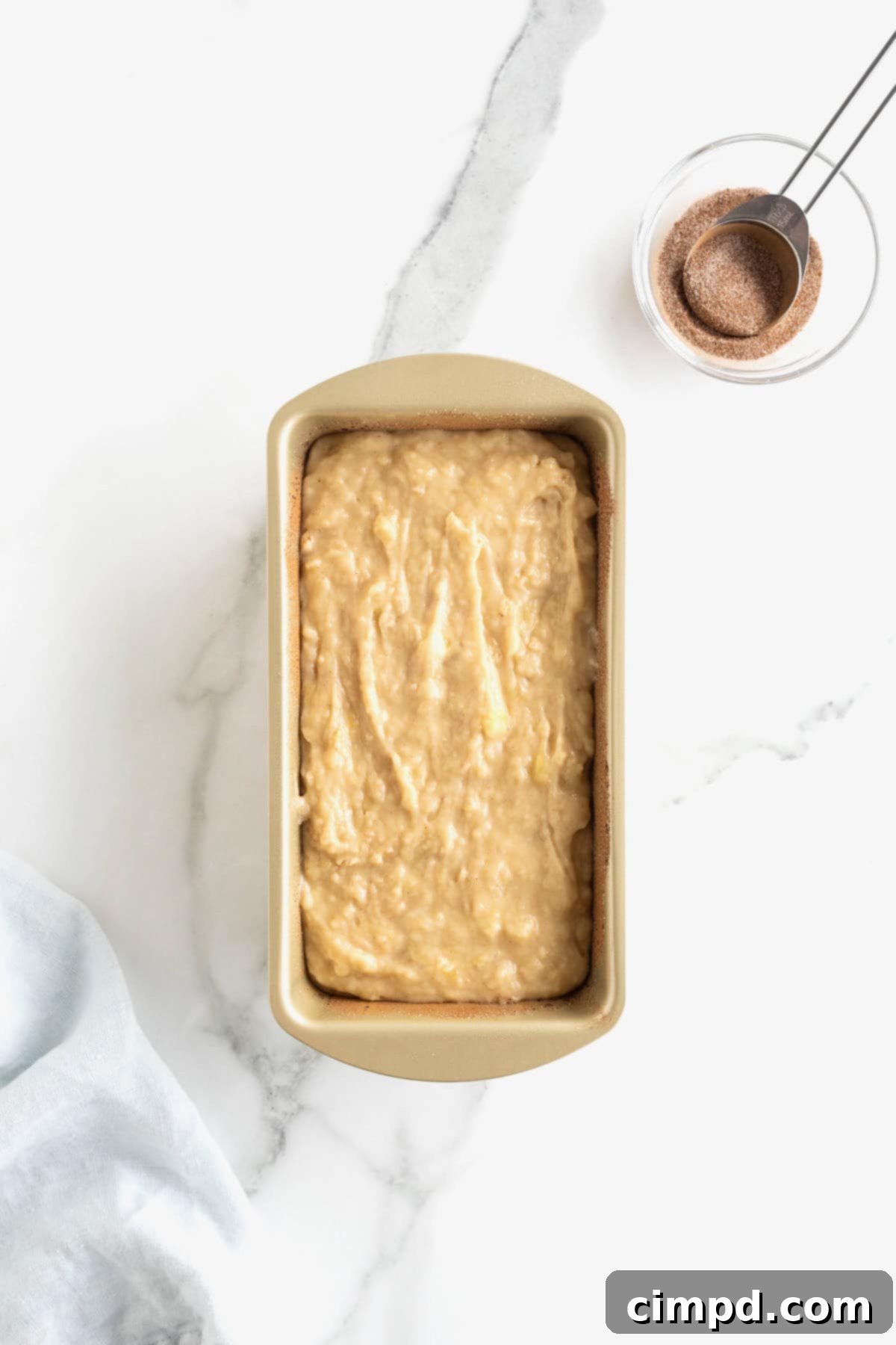 A gleaming gold-toned loaf pan, perfectly filled with a smooth, light brown banana bread batter, sits invitingly on a white marble counter. To the top right, a small white dish holds a tempting mound of cinnamon sugar, ready for the next step.