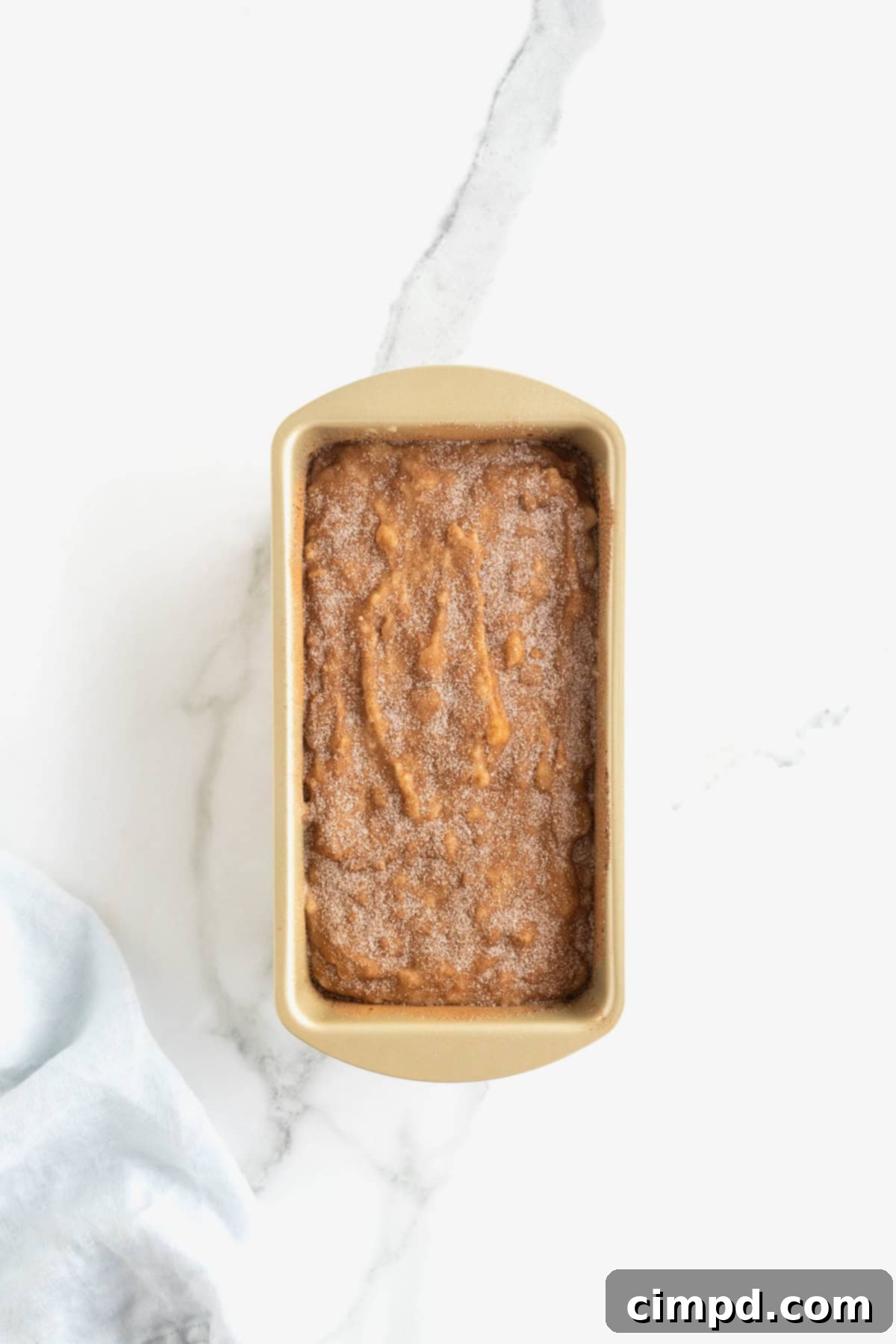 A gold-toned loaf pan, now filled with banana bread batter, is seen with a generous and even sprinkle of cinnamon sugar across its surface, promising a delicious, crackly crust upon baking.
