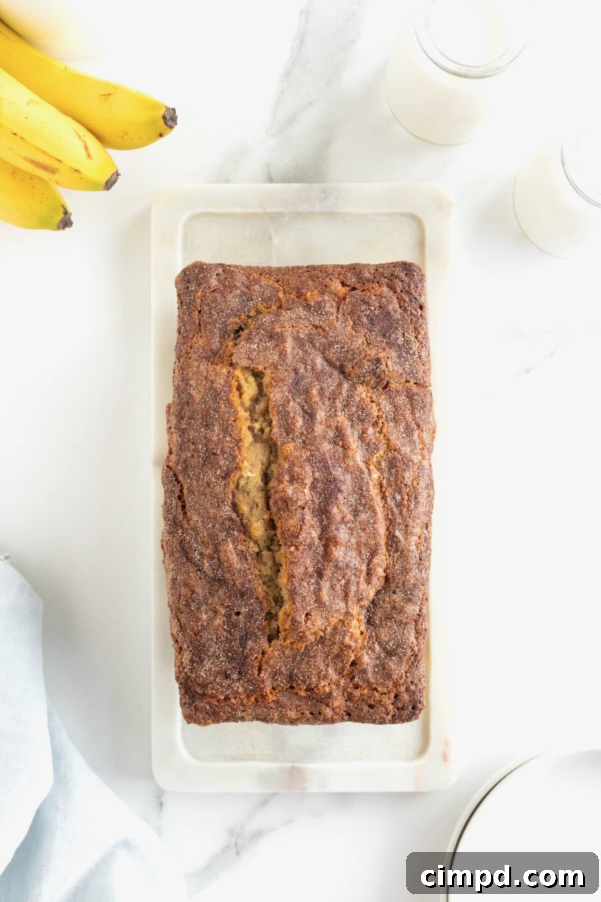 A perfectly baked loaf of golden-brown Cinnamon Banana Bread, fresh from the oven, resting on a white stone cutting board on a white marble counter, its cinnamon sugar crust glistening.