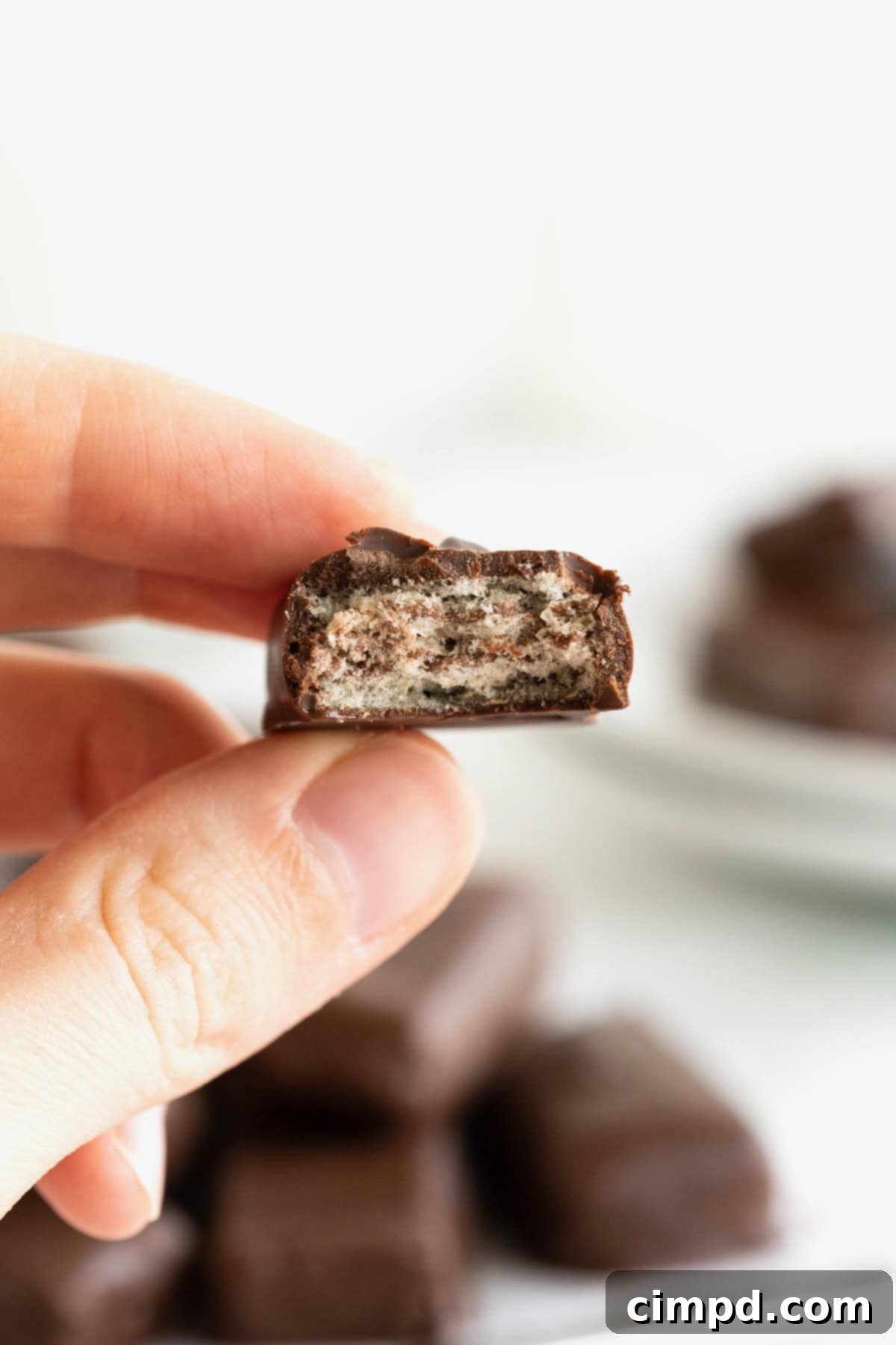 Quick Homemade KitKat Bites 5 A hand holding a homemade KitKat bite, perfectly coated in chocolate, above a stack of similar bites on a parchment-lined rack.