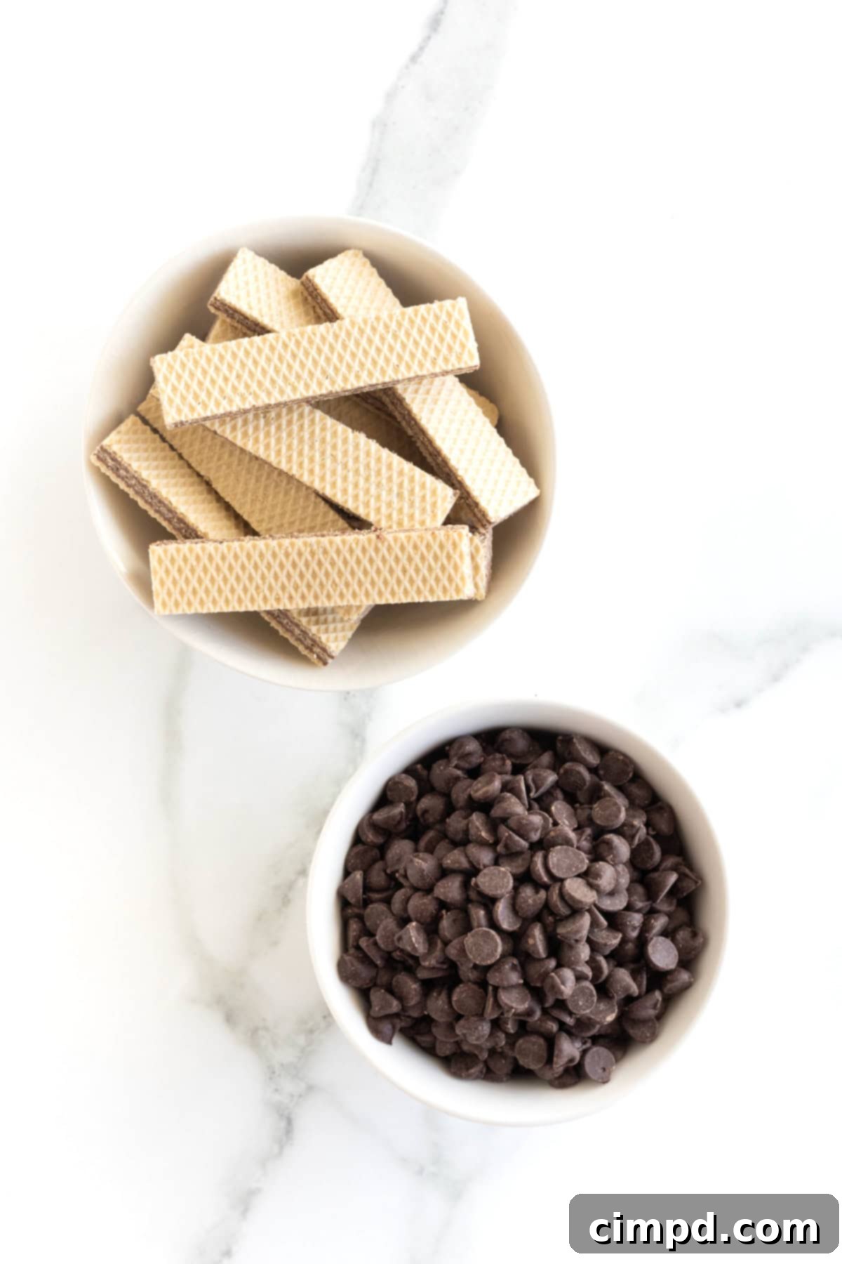 Quick Homemade KitKat Bites 6 A rustic shot of a white bowl overflowing with chocolate crème filled wafer cookies, positioned next to a smaller white bowl brimming with mini semi-sweet chocolate chips, all resting on a elegant white marble countertop, ready for baking.