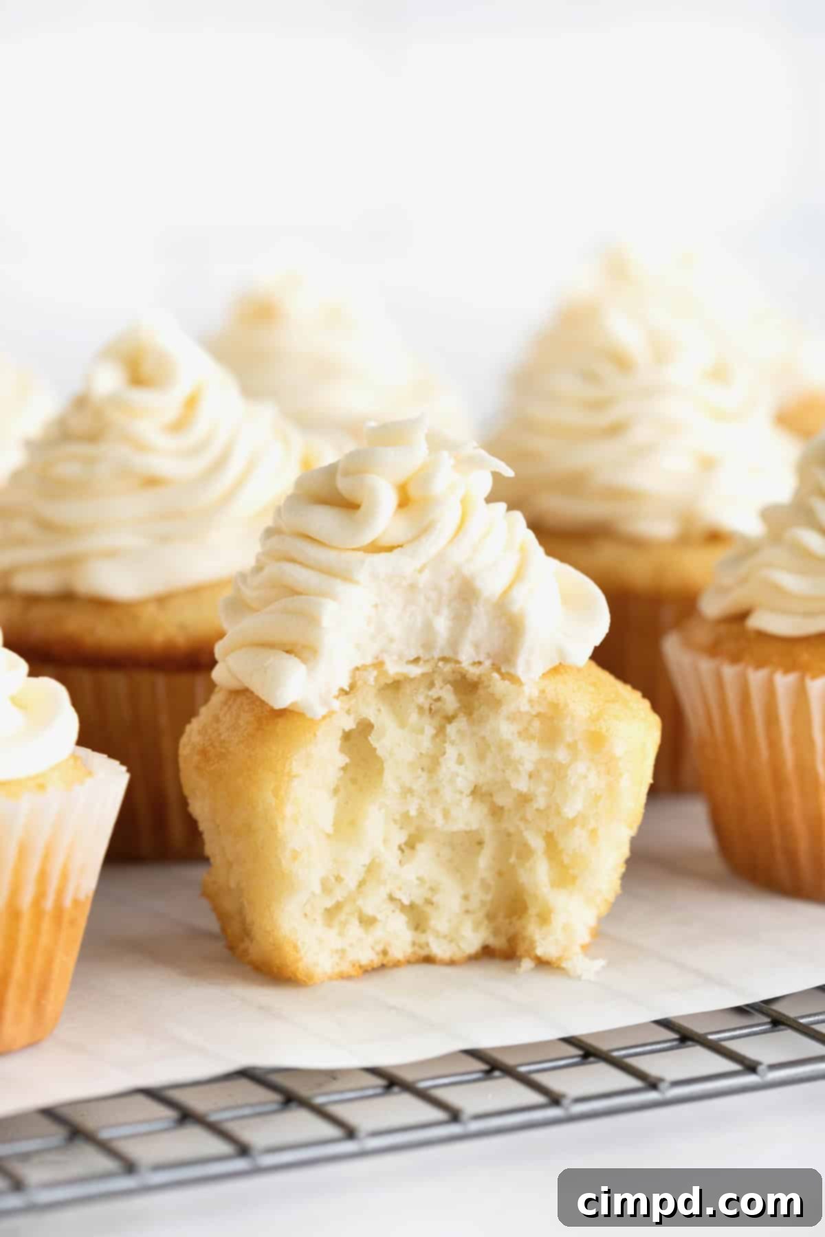 Foolproof Buttercream Frosting 3 Six vanilla cupcakes piped with buttercream frosting sitting on a parchment lined cooling rack. The center cupcake has a bite bit out it.
