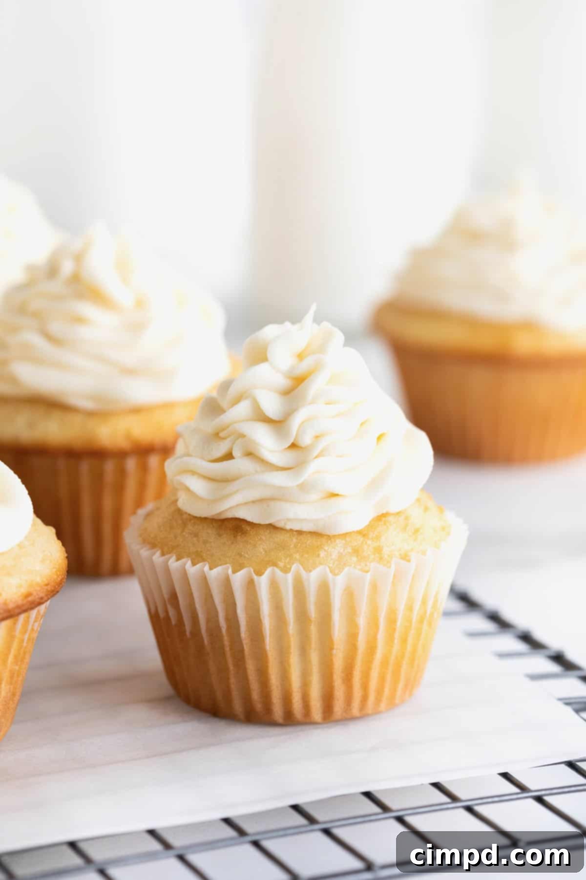 Foolproof Buttercream Frosting 4 Vanilla cupcakes piped with buttercream frosting sitting on a parchment lined cooling rack.