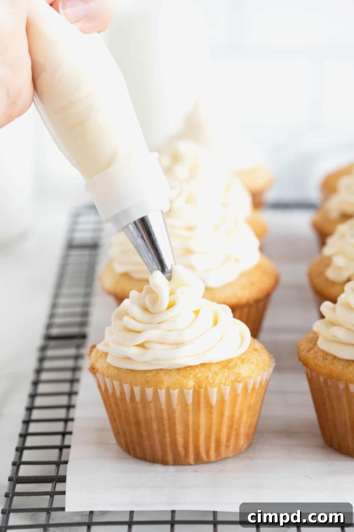 Foolproof Buttercream Frosting 9 Buttercream frosting being piped onto a vanilla cupcake on a parchment lined cooling rack.