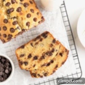 Decadent Chocolate Chip Pound Cake 11 A slice of chocolate chip pound cake in front of a loaf of pound cake on a parchment lined cooling rack. A small white dish of chocolate chips sits to the bottom left of the photo.