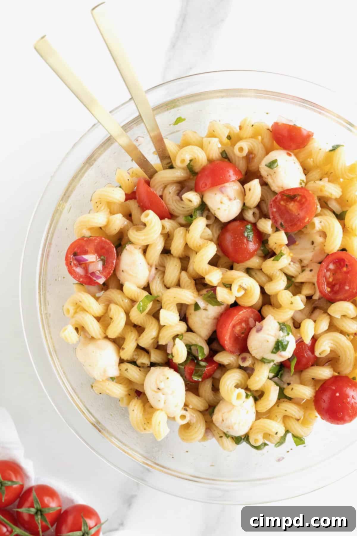 A large glass serving bowl brimming with Caprese pasta salad, featuring halved bright red cherry tomatoes, pearl-sized mozzarella balls, and corkscrew pasta. A rustic wooden spoon is gently placed in the bowl, ready for serving.