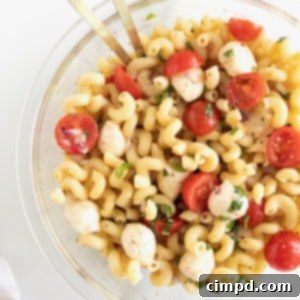 A large glass serving bowl of capers pasta salad with halved cheery tomatoes, bite sized mozzarella balls and corkscrew pasta.
