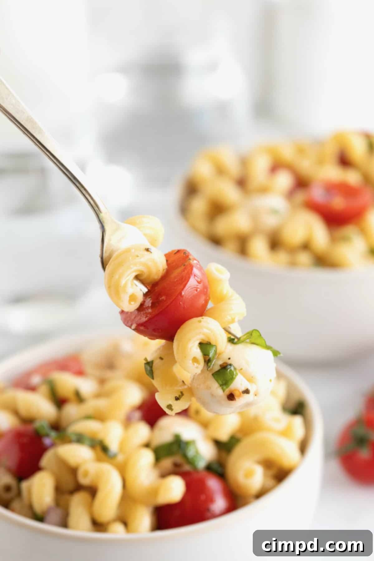 A fork holding a perfect bite of Caprese pasta salad, showcasing a piece of mozzarella, a tomato half, and pasta, with a blurred bowl of the full salad in the background.
