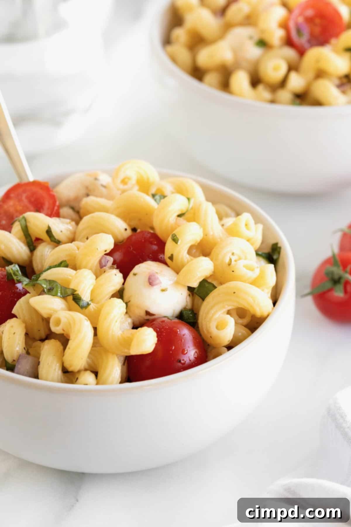 A pristine white bowl filled with Caprese pasta salad rests on a white marble counter, with a metal fork poised within the salad.