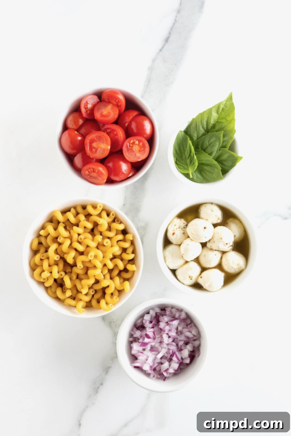 Various ingredients for Caprese pasta salad, neatly arranged in small white glass dishes on a white marble counter, including pasta, cherry tomatoes, red onion, mozzarella, and fresh basil.