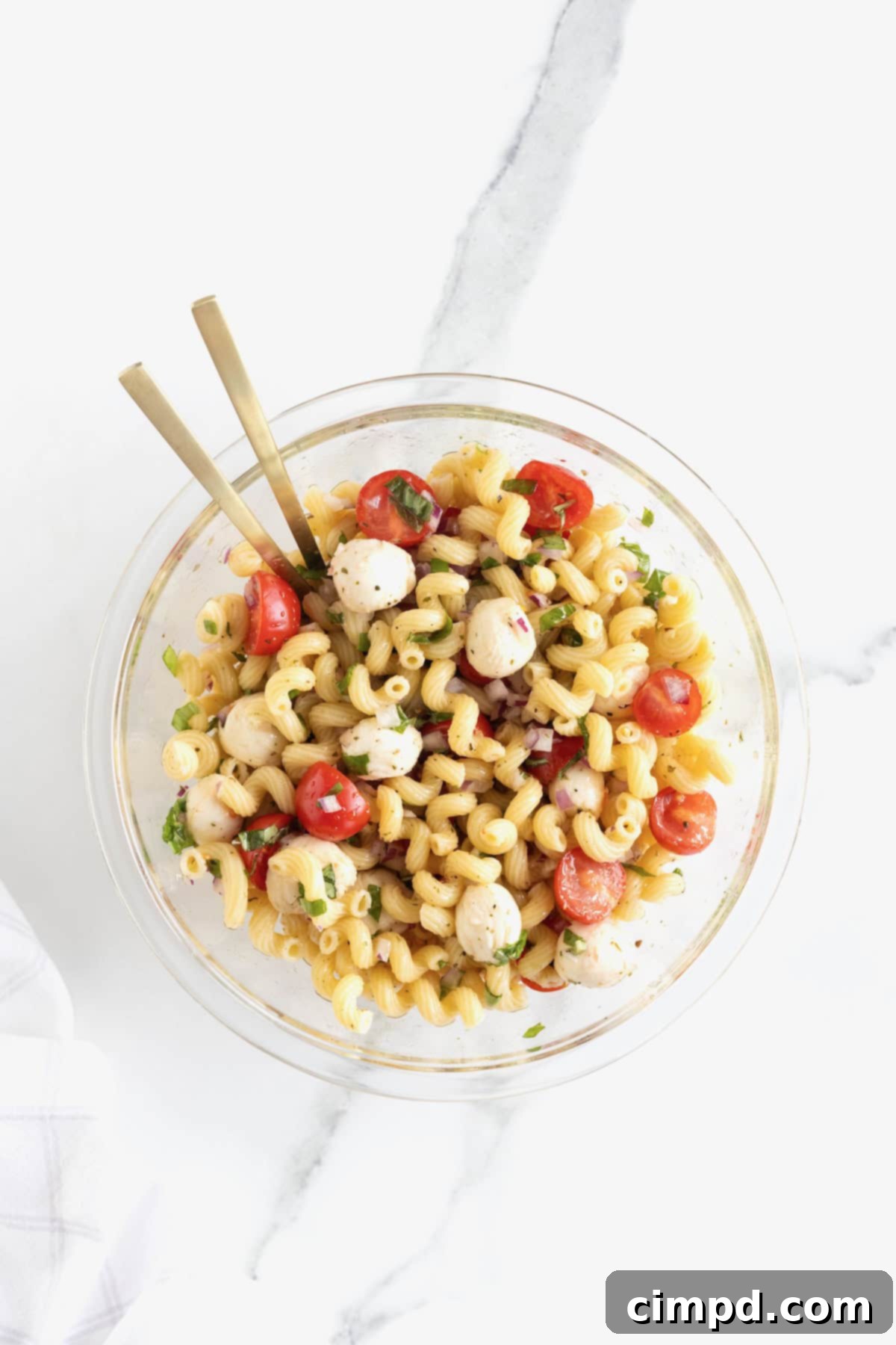 A large clear glass serving bowl filled with a colorful Caprese pasta salad, highlighting the cherry tomatoes, mozzarella balls, and corkscrew pasta, presented on a light background.