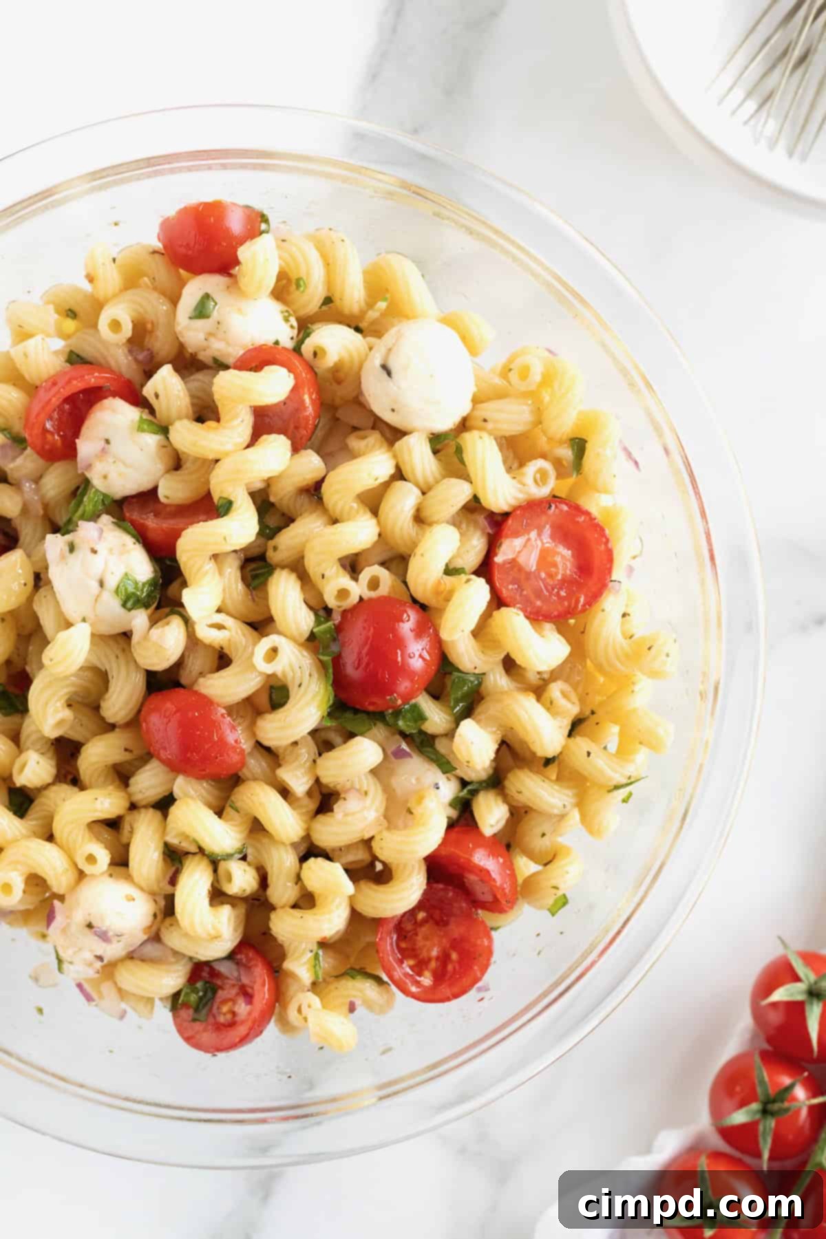 A large glass serving bowl of Caprese pasta salad with halved cherry tomatoes, bite-sized mozzarella balls, and corkscrew pasta, captured from a slightly elevated angle.