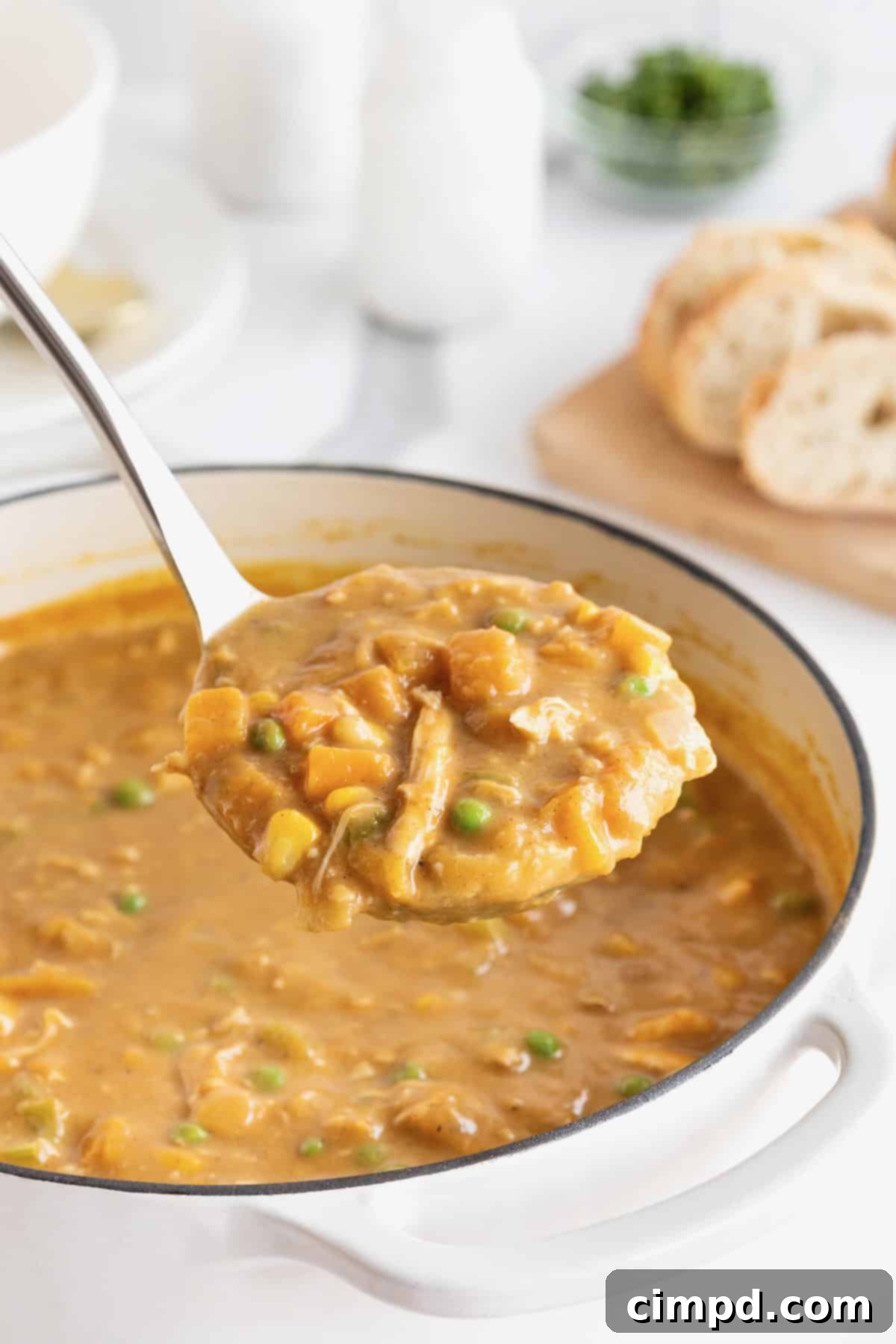 A ladle gracefully dishes out a serving of golden Pumpkin Pot Pie Soup from a pristine white Dutch oven, resting on a gleaming white marble countertop.