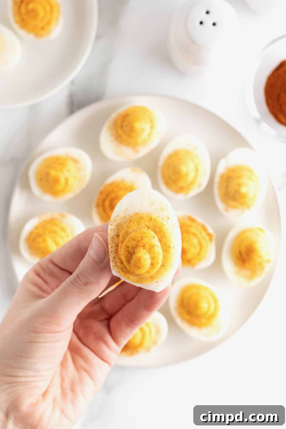 Devilish Delights 4 A hand reaches for a deviled egg from a generous white serving platter, brimming with more deviled eggs, all resting on a white marble counter.
