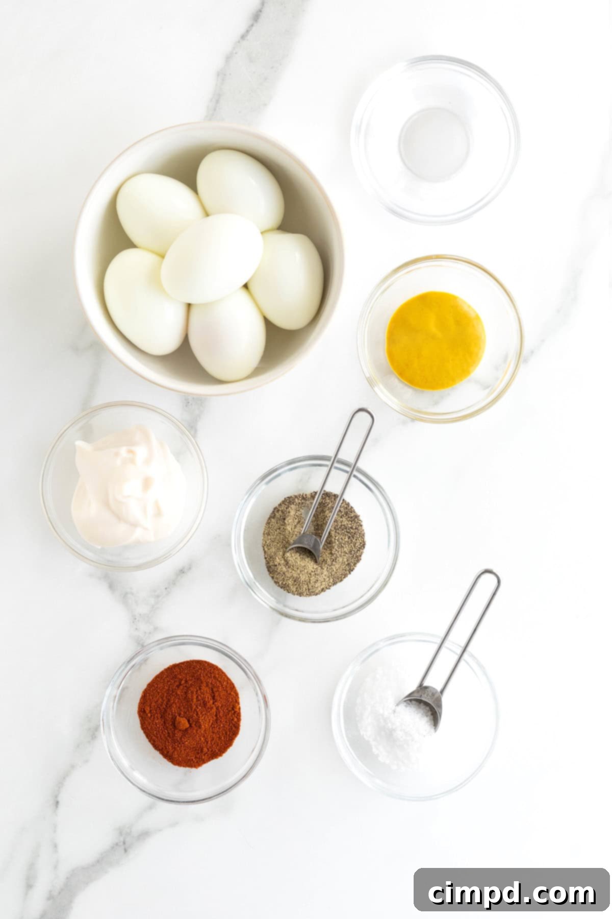 Devilish Delights 5 Small glass dishes filled with the fresh ingredients needed to make deviled eggs, including mayonnaise, mustard, vinegar, salt, pepper, and paprika, arranged on a white marble counter.