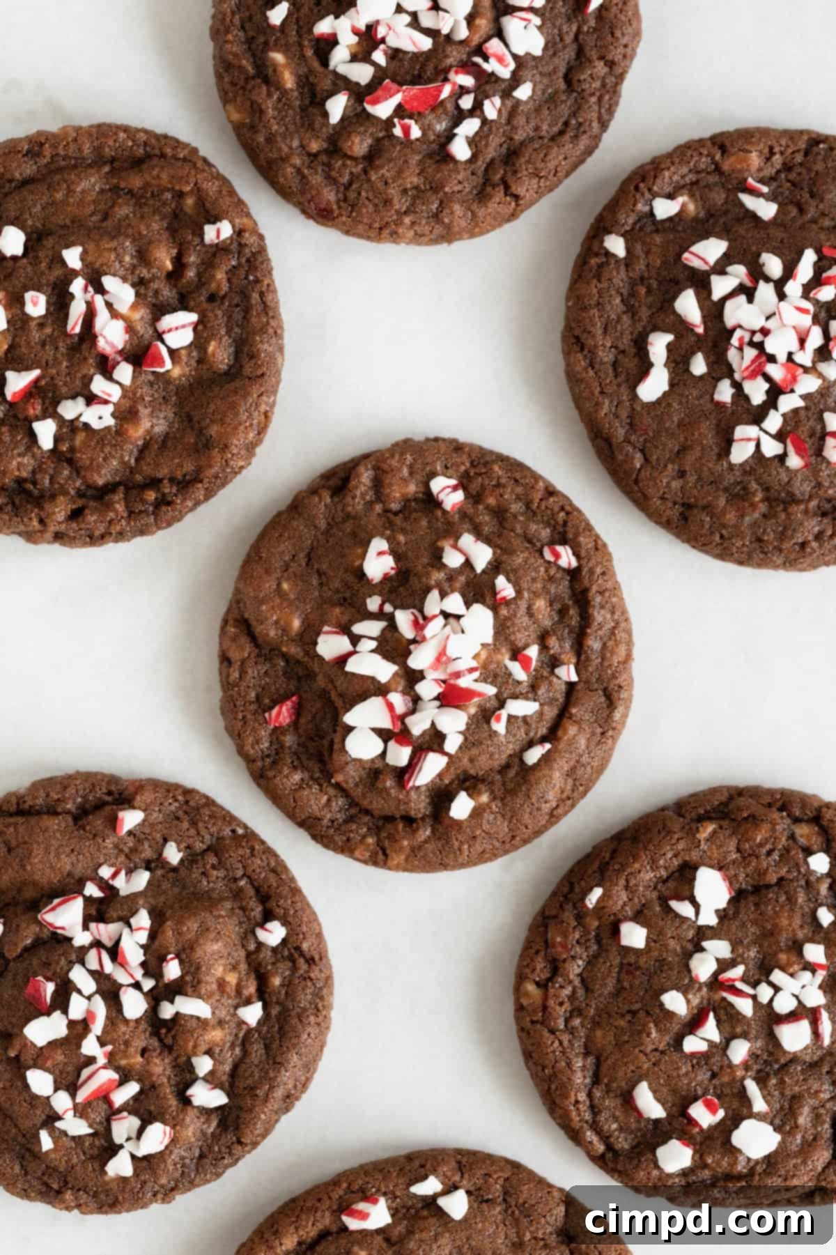 Seven rich chocolate cookies, generously topped with vibrant peppermint bits, resting on a rustic parchment paper surface.