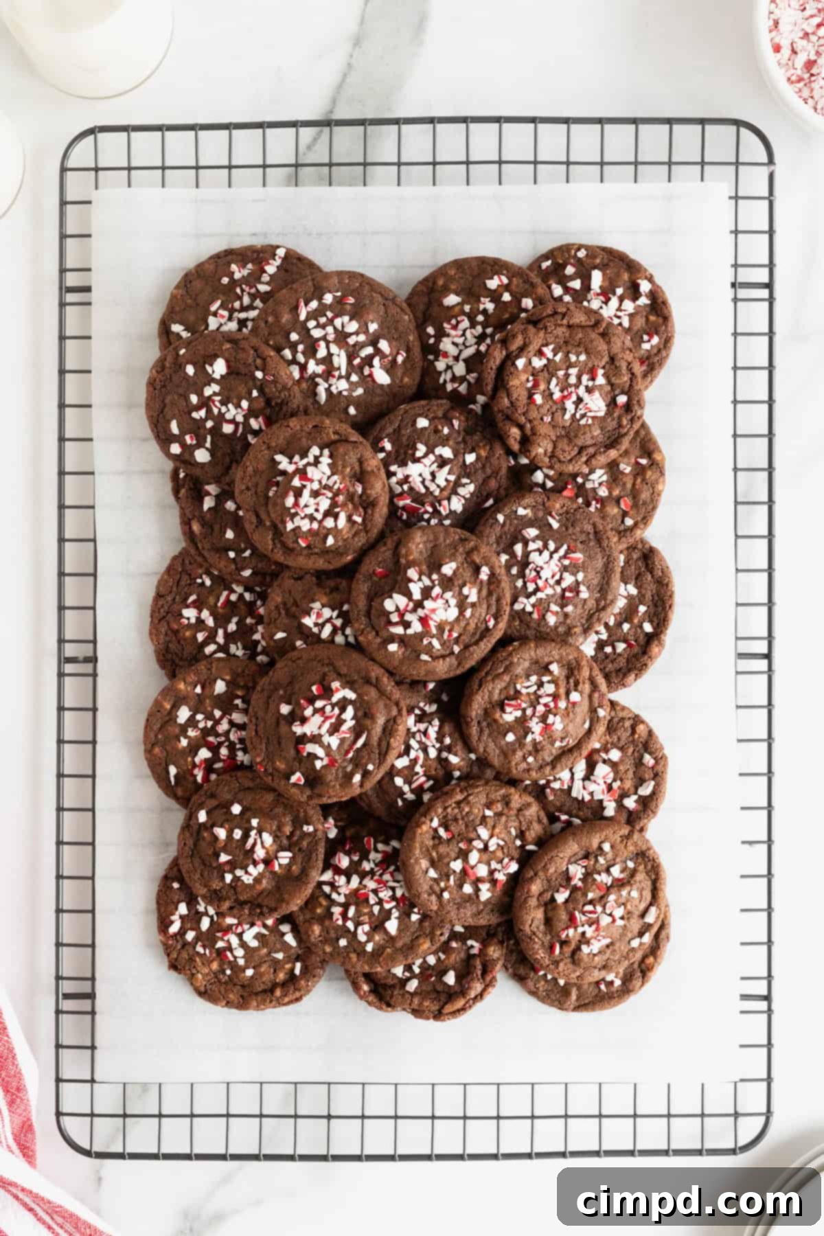 A delightful pile of rich chocolate cookies, generously adorned with vibrant peppermint bits and delicate white chocolate drizzle, cooling on a parchment-lined rack.