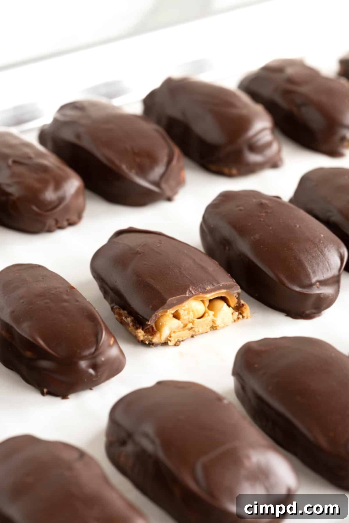 Chewy Homemade Snickers Treats 3 Perfectly arranged rows of homemade Snickers bites on a parchment-lined aluminum baking sheet, with one piece ready to be enjoyed.