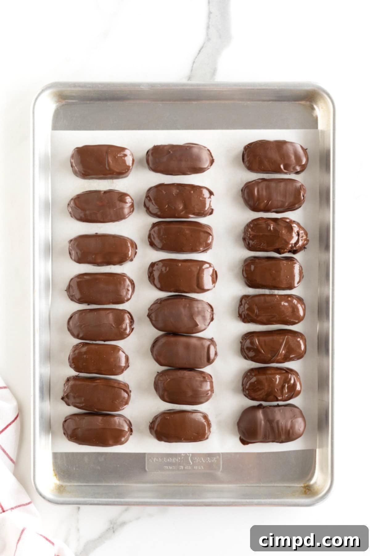 Chewy Homemade Snickers Treats 10 Rows of freshly coated homemade Snickers bites neatly arranged on a parchment-lined aluminum baking sheet, allowing the chocolate to set.