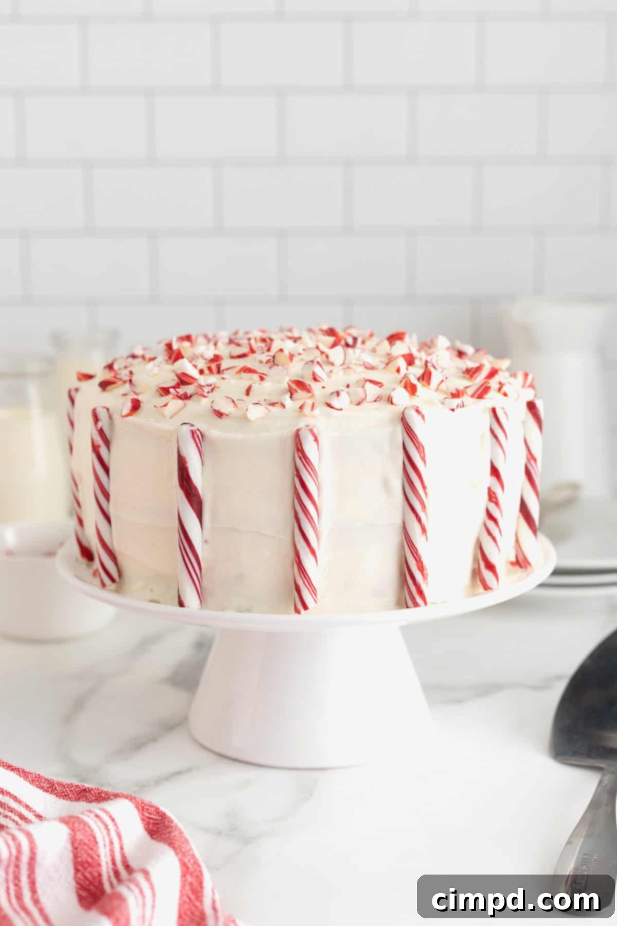 Peppermint Swirl Cake 13 A festive two-layer cake showcasing pristine white frosting, beautifully garnished with whole candy canes and delightful crushed peppermint pieces, elegantly displayed on a white cake stand.