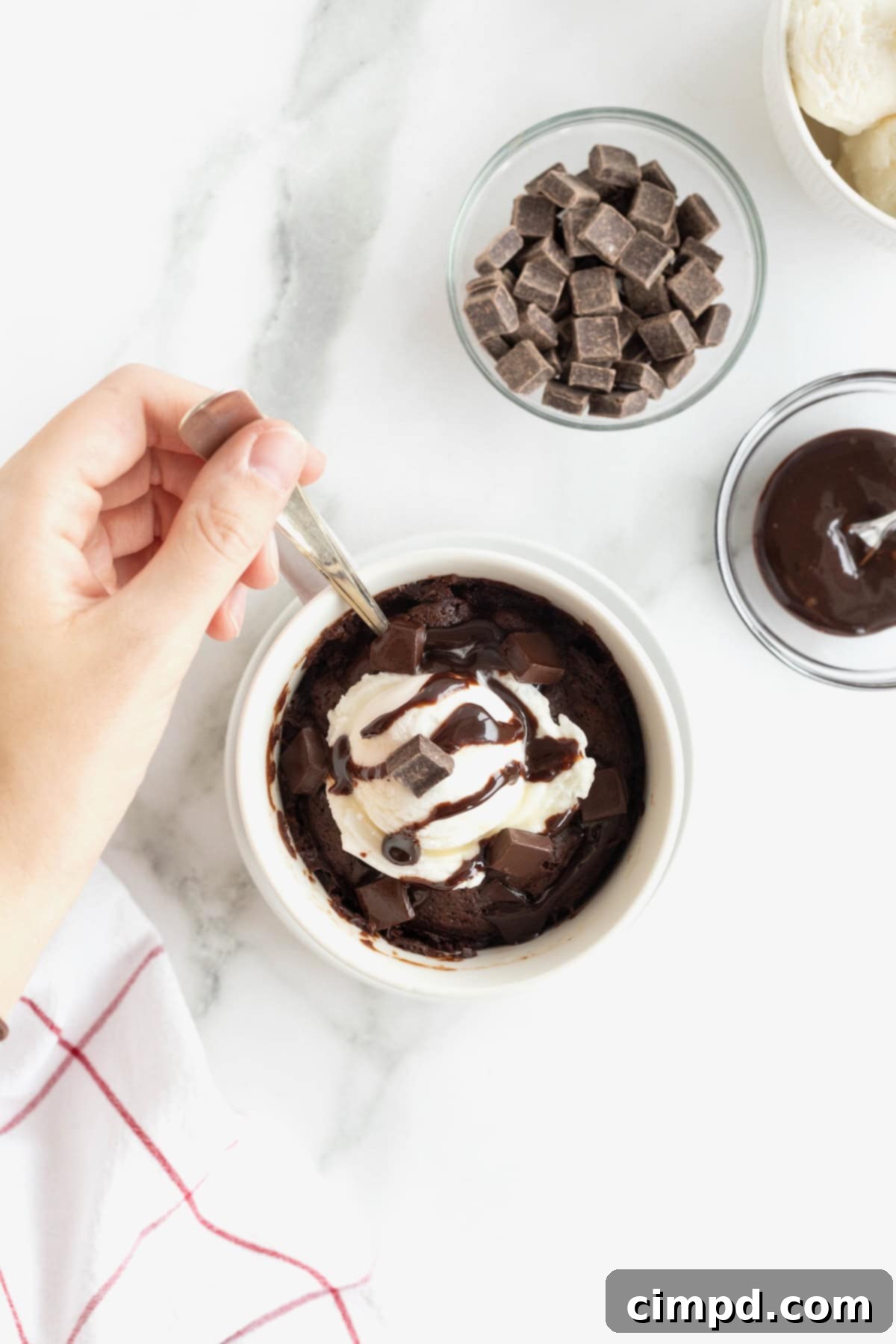 Speedy Chocolate Mug Brownie 4 A perfectly cooked microwave brownie served in a white mug, elegantly placed on a white saucer. It's crowned with a generous scoop of creamy vanilla ice cream and a luscious chocolate syrup drizzle.