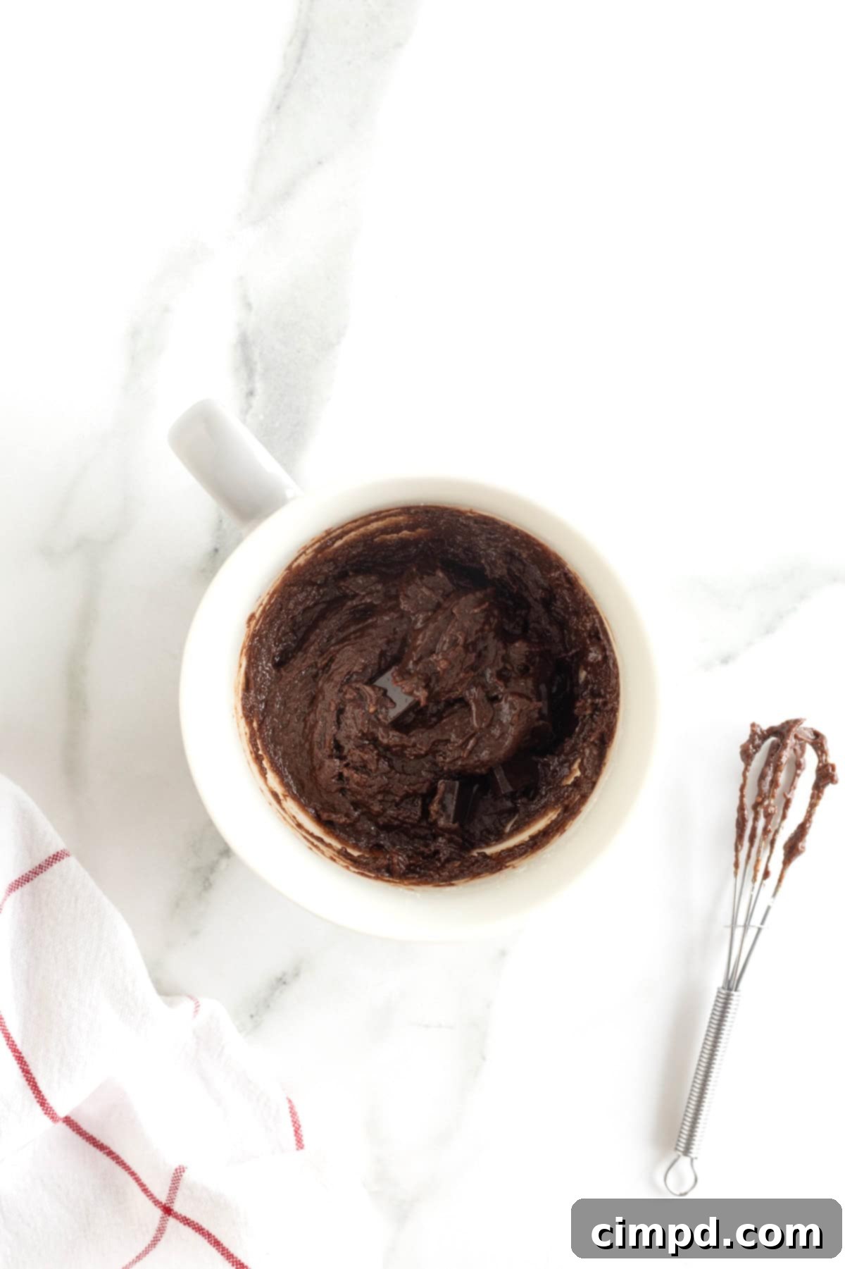 Speedy Chocolate Mug Brownie 6 Rich chocolate brownie batter filling a white mug, resting on a white marble counter. A mini whisk, covered in chocolate, lies casually to the bottom right.