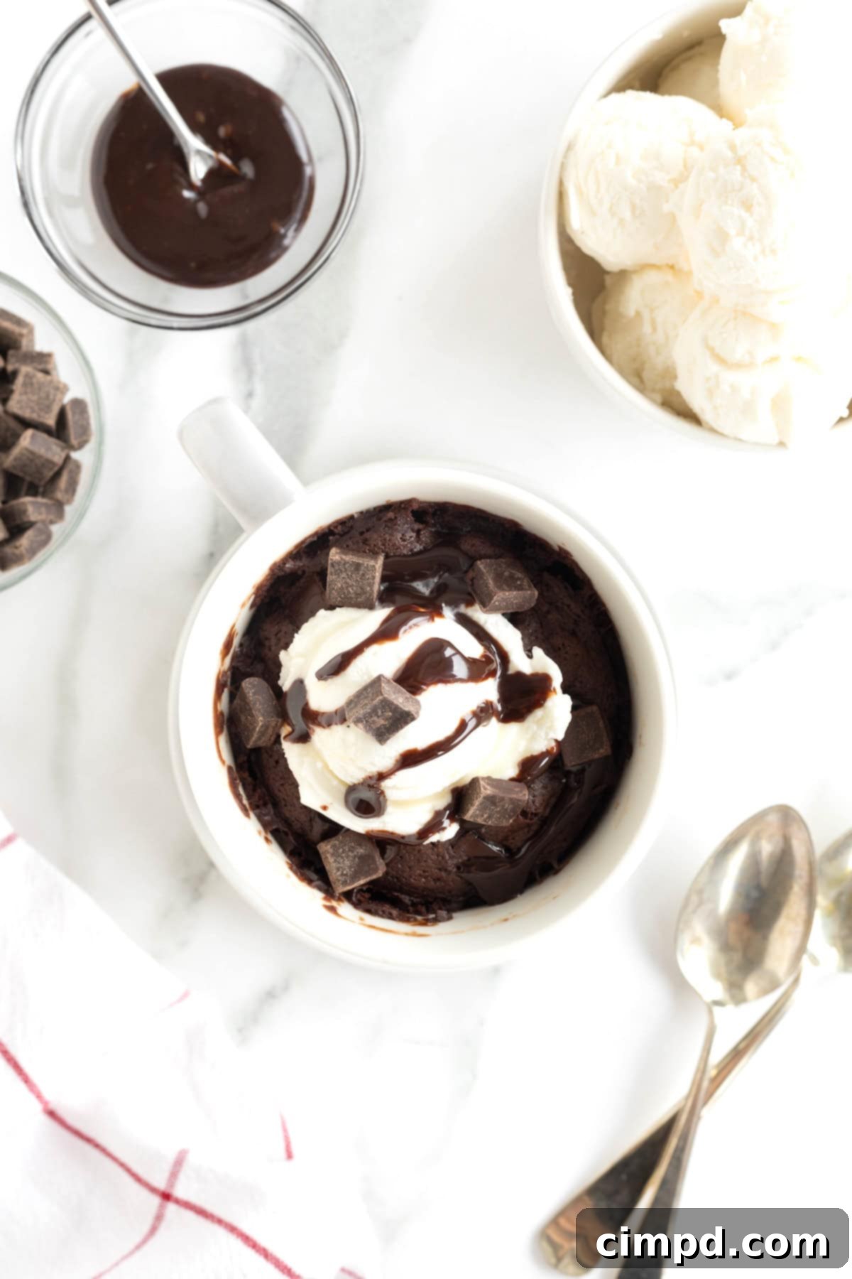 Speedy Chocolate Mug Brownie 8 A decadent microwave brownie in a white mug, lavishly topped with a scoop of vanilla ice cream, a rich chocolate sauce drizzle, and extra chocolate chunks, all presented on a white saucer.