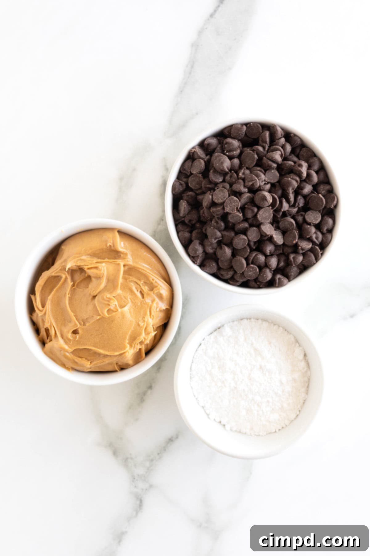 Three Ingredient Peanut Butter Cups 6 All the necessary ingredients to make peanut butter cups artfully arranged in small white dishes on a pristine white marble countertop.