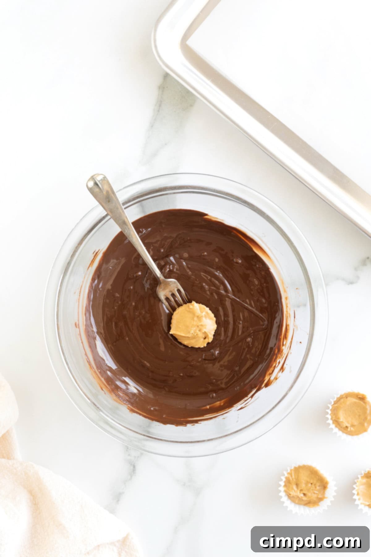 Three Ingredient Peanut Butter Cups 9 A close-up shot of a peanut butter cup being gracefully dipped into a bowl of rich, melted chocolate with a fork, poised over a white marble countertop.