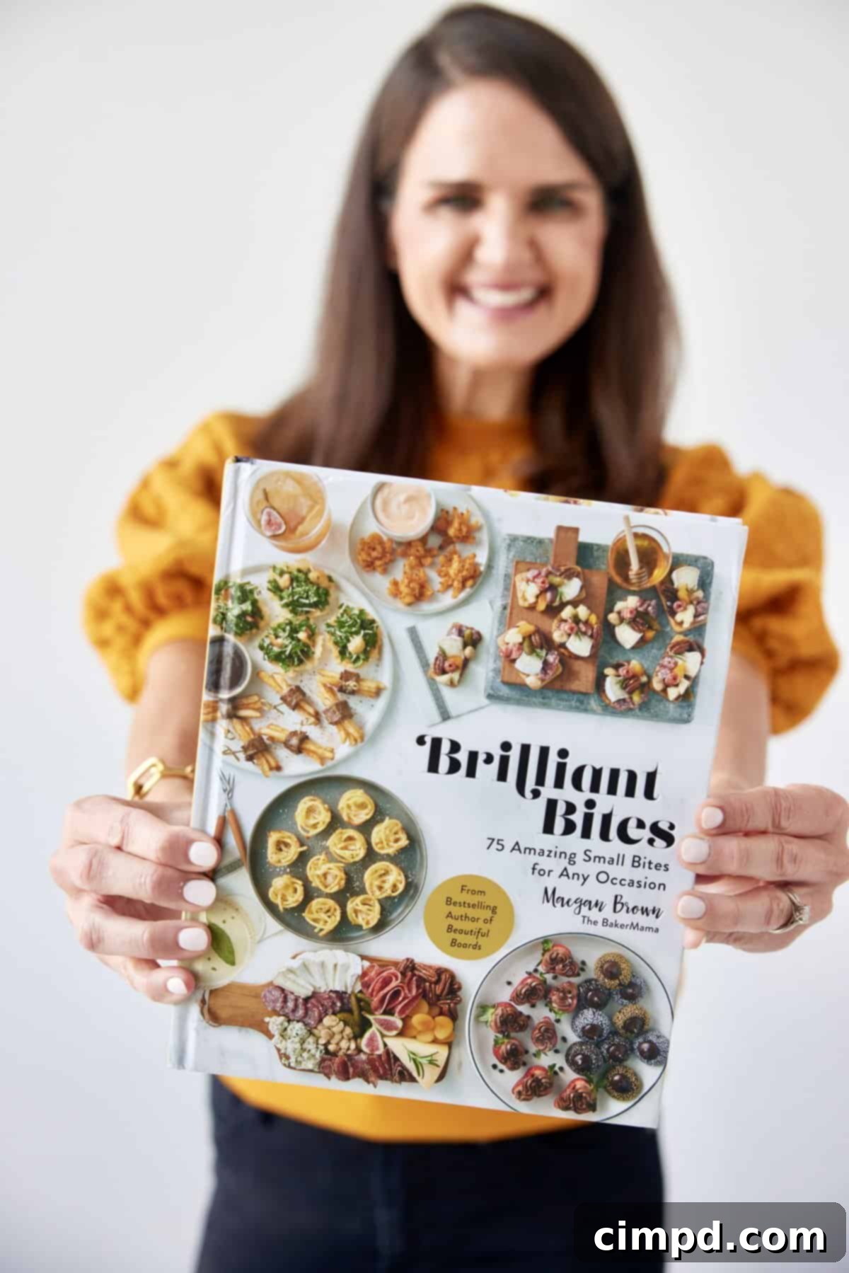 Maegan Brown wearing a marigold yellow top holding her cookbook, Brilliant Bites, out in front of her.