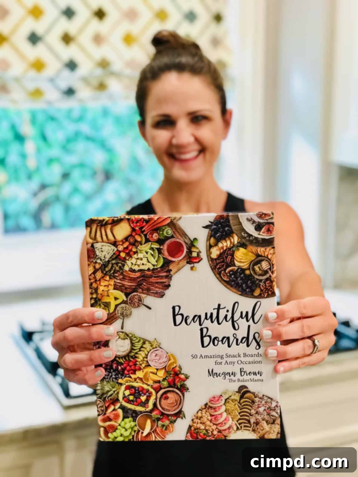 Maegan Brown, author of Beautiful Boards, holding a copy of her popular cookbook with a warm smile, highlighting her accomplishment.